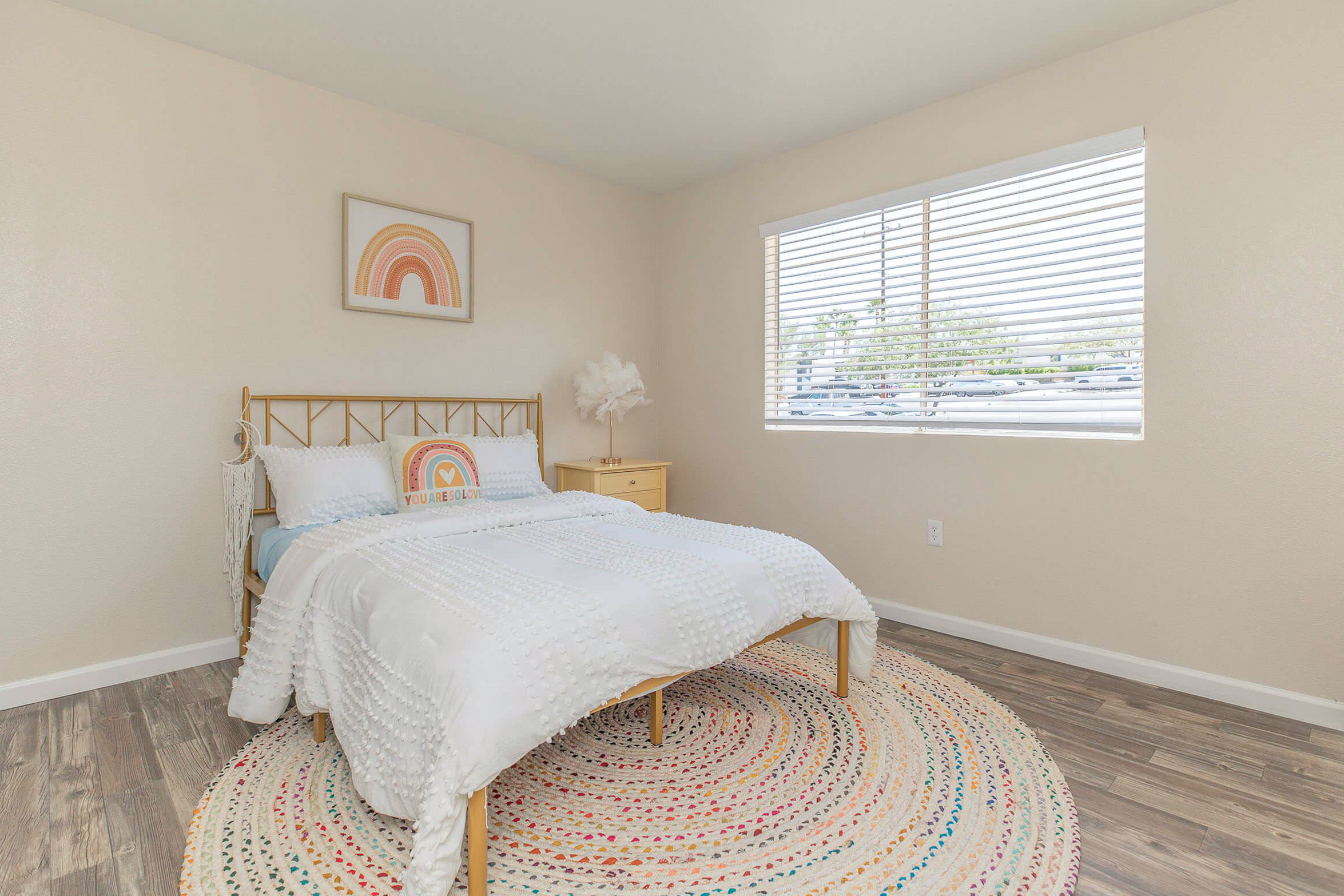 A bright and airy bedroom featuring a gold metal bed with white bedding, a colorful rainbow artwork above the bed, a round braided rug on the floor, and a nightstand. Natural light filters through the window with white blinds, illuminating the neutral-colored walls and wood-like flooring.