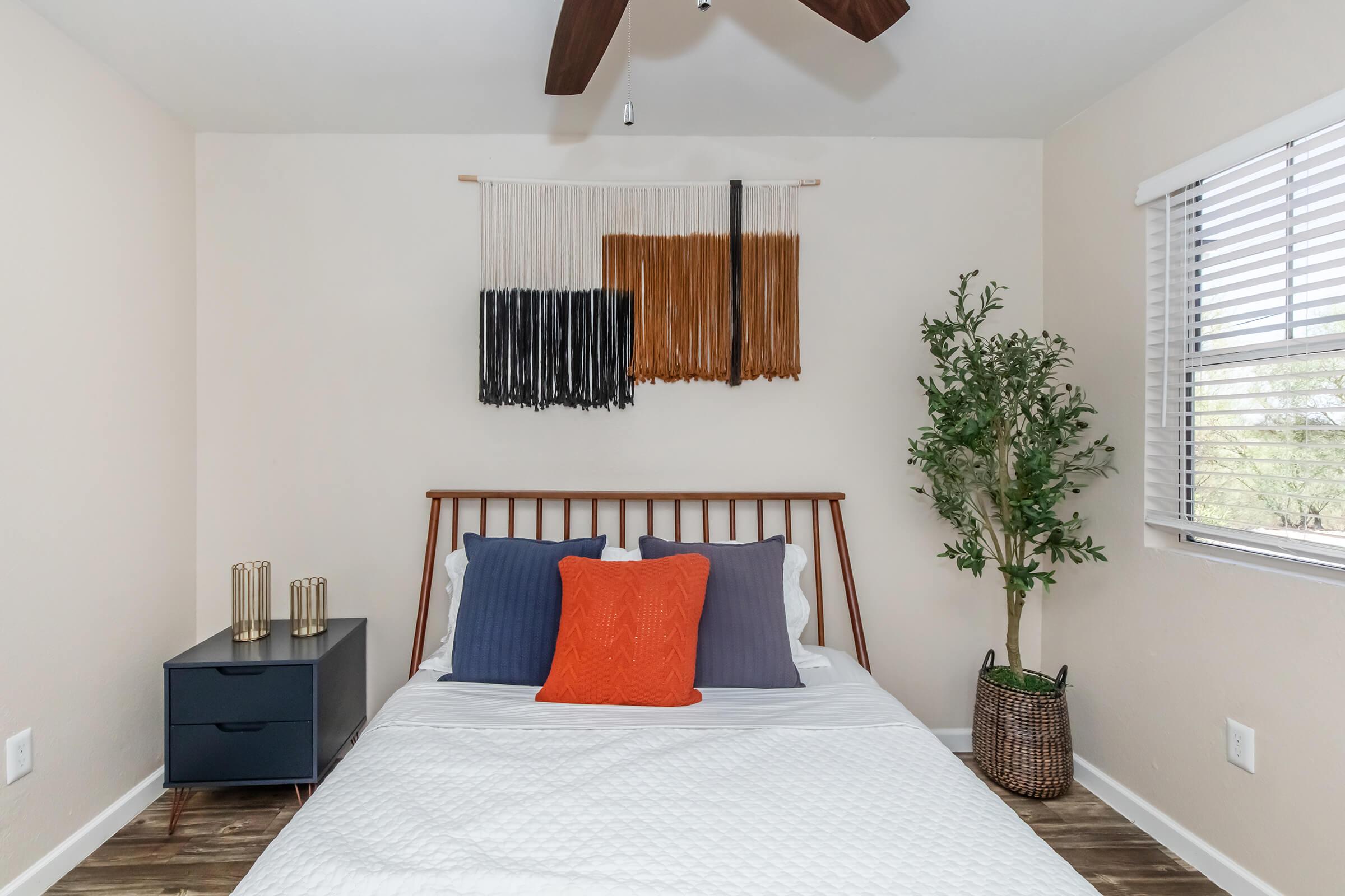 A cozy bedroom with a modern design featuring a wooden bed frame with white bedding and decorative pillows in blue and orange. A nightstand holds three metallic vases, and a potted plant adds greenery. A wall hanging in neutral tones complements the space, and there are window blinds that allow natural light.