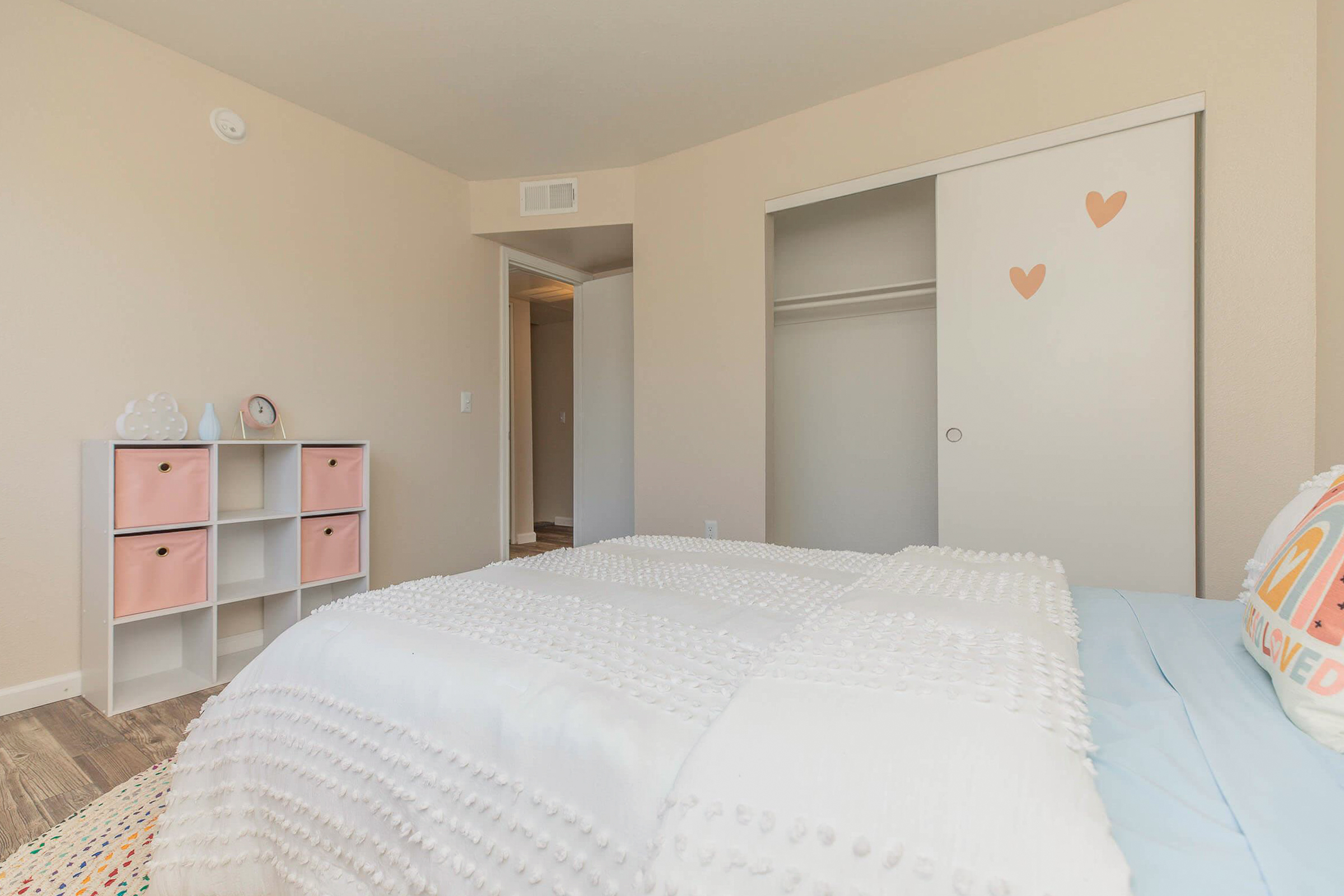 A softly decorated bedroom featuring a light-colored bedspread, a bookshelf with pink and white accents, and a closet with heart-shaped decals. The walls are a light tan color, and the flooring is wood-like. A doorway leads to another room in the background.