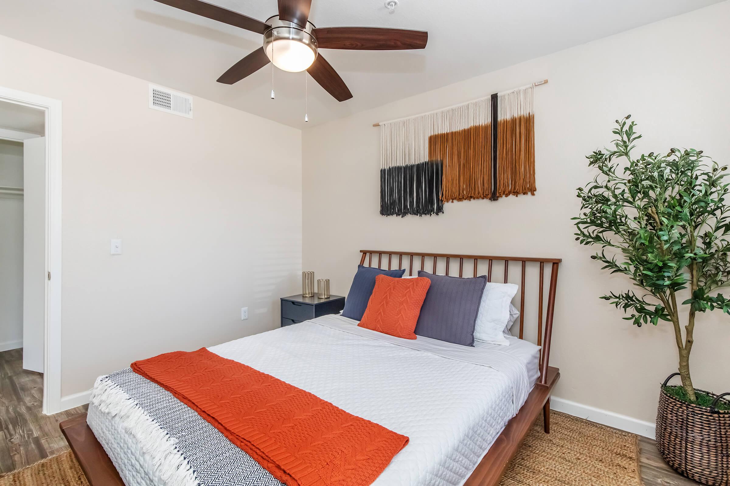 A cozy bedroom featuring a wooden bed with white and orange bedding, decorative pillows, and a ceiling fan. The walls are adorned with a textile wall hanging in neutral tones, and a small green plant is placed beside a nightstand. Natural light brightens the space, creating a warm atmosphere.