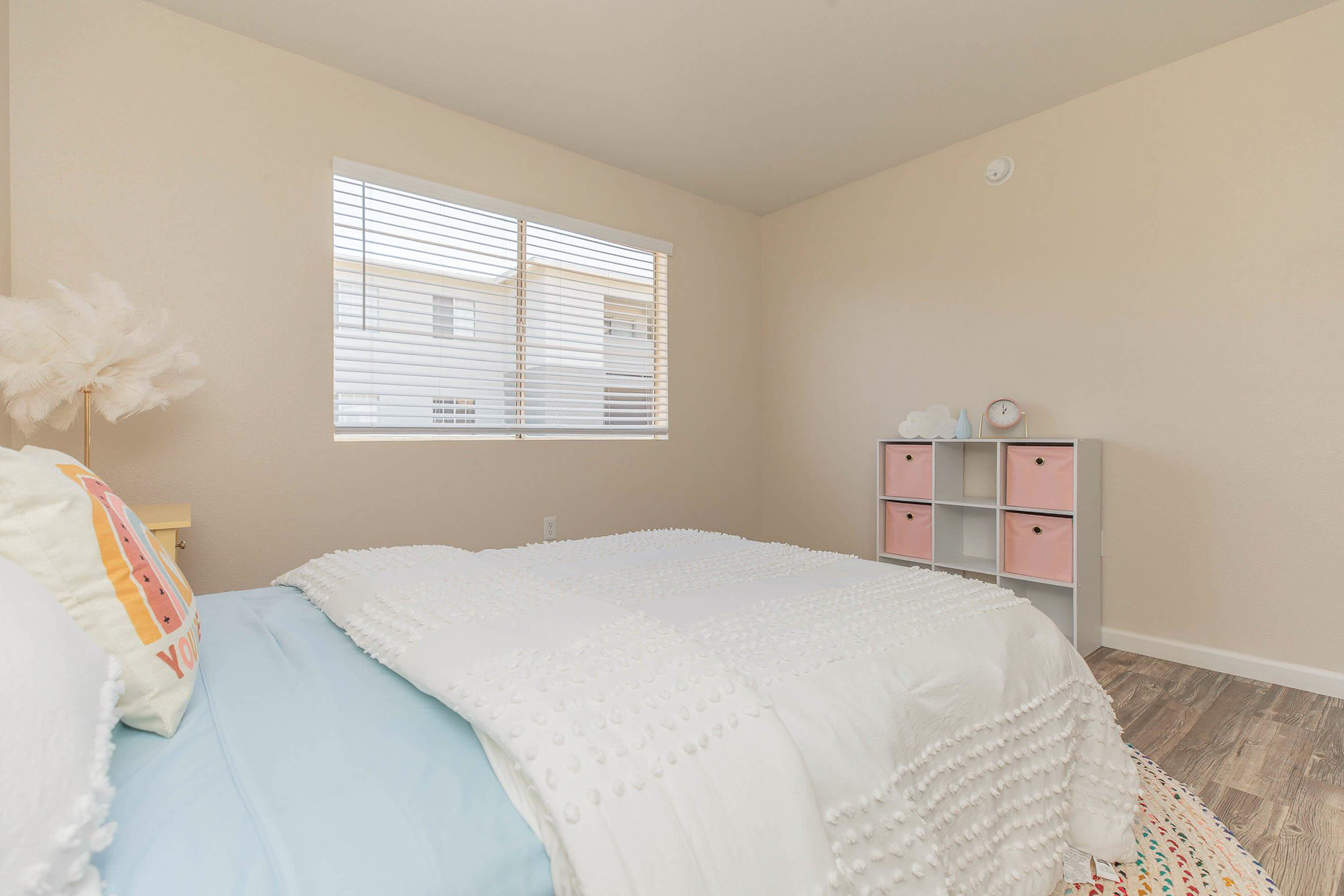 A cozy bedroom featuring a bed with a textured white blanket and a pastel blue sheet. There's a small bedside table with a decorative lamp and a fluffy pink decor item. A window with blinds allows soft light to enter, and a storage unit with pink bins is positioned against the wall. The walls are painted a neutral color.