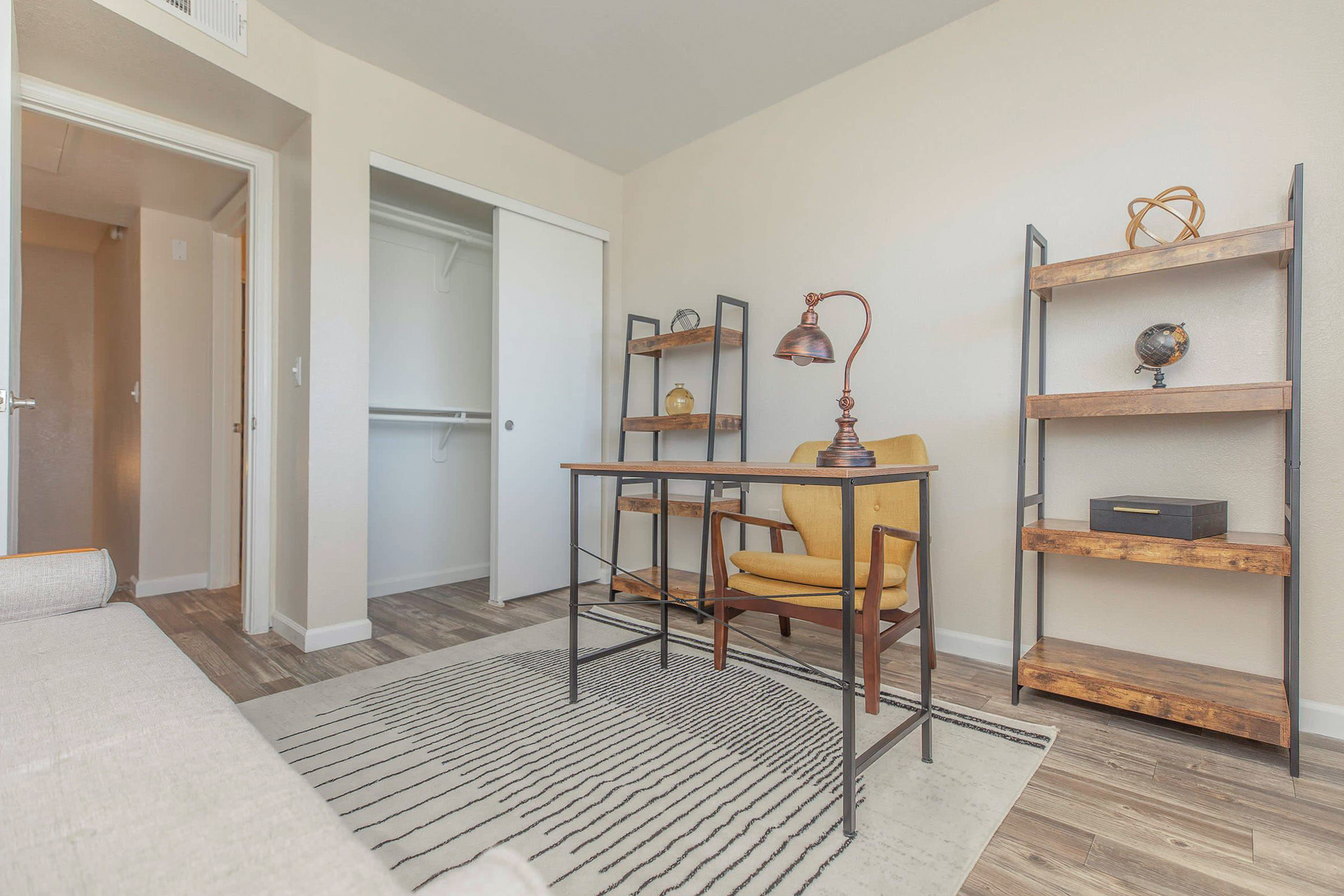 A modern and tidy room featuring a wooden desk with a vintage lamp, an upholstered yellow chair, and two bookshelves with decorative items. The floor is covered with a patterned rug, and there is a closet door visible on the left. Natural light brightens the space, creating a warm and inviting atmosphere.
