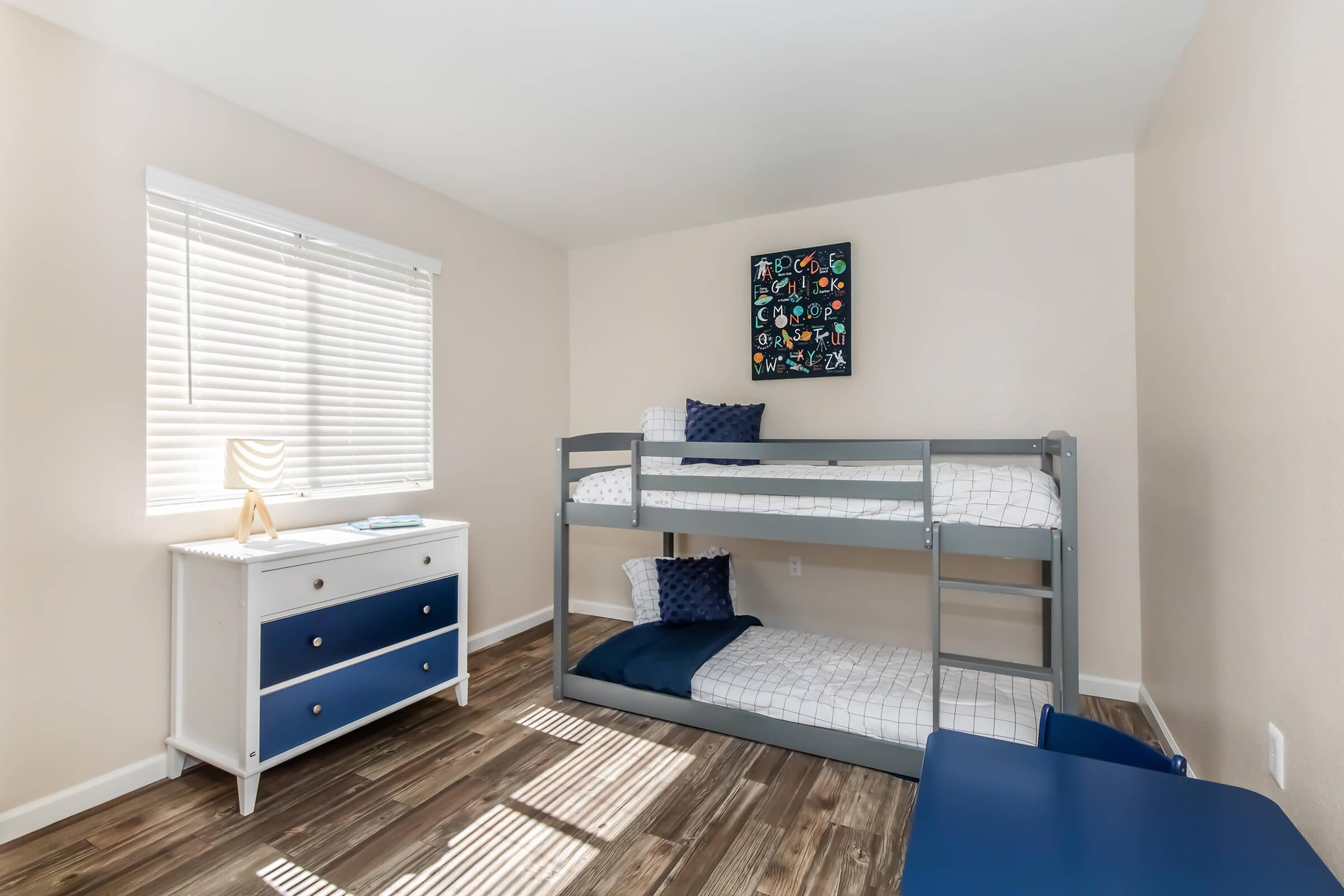 A cozy bedroom featuring a gray bunk bed with white bedding, a blue accent pillow, and a blue bedside table. Light streams in through a window with white blinds, and a colorful wall art piece hangs above the bed. The floor is wooden, creating a warm atmosphere.
