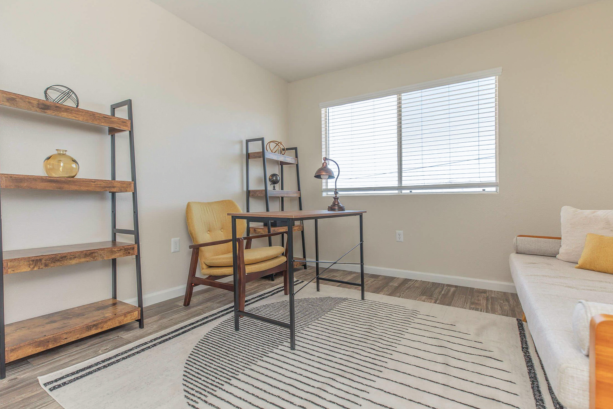 A bright, minimalistic room featuring a wooden desk with a lamp, a retro-style armchair in mustard yellow, and a shelf with decorative items. The floor is covered with a geometric-patterned rug, and large windows allow natural light to brighten the space. The walls are neutral, enhancing the airy feel.