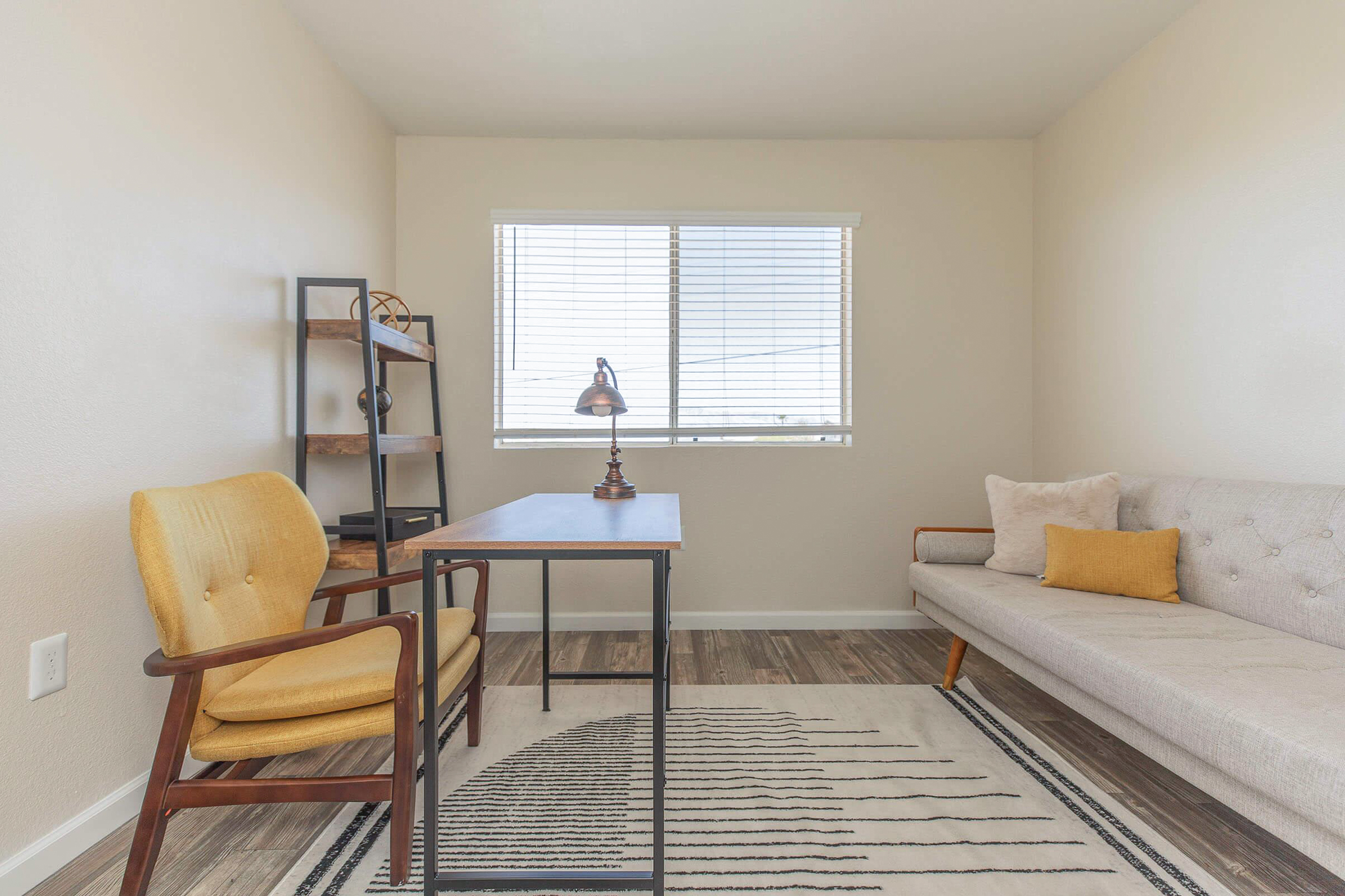 A bright, minimalist home office featuring a small desk with a lamp, a comfortable yellow chair, and a modern gray sofa. There's a decorative shelf against the wall and a window with blinds allowing natural light to brighten the space. A patterned area rug adds texture to the floor.