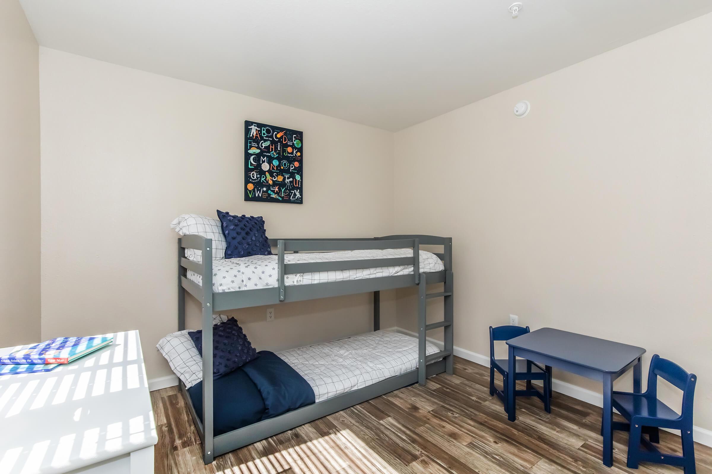 A bedroom featuring a gray bunk bed with white bedding, a blue throw pillow on the top bunk, and a small blue table with two chairs. The walls are a light beige color, and there is a colorful framed artwork on the wall above the bed. Sunlight is streaming through a window, creating striped shadows on the floor.
