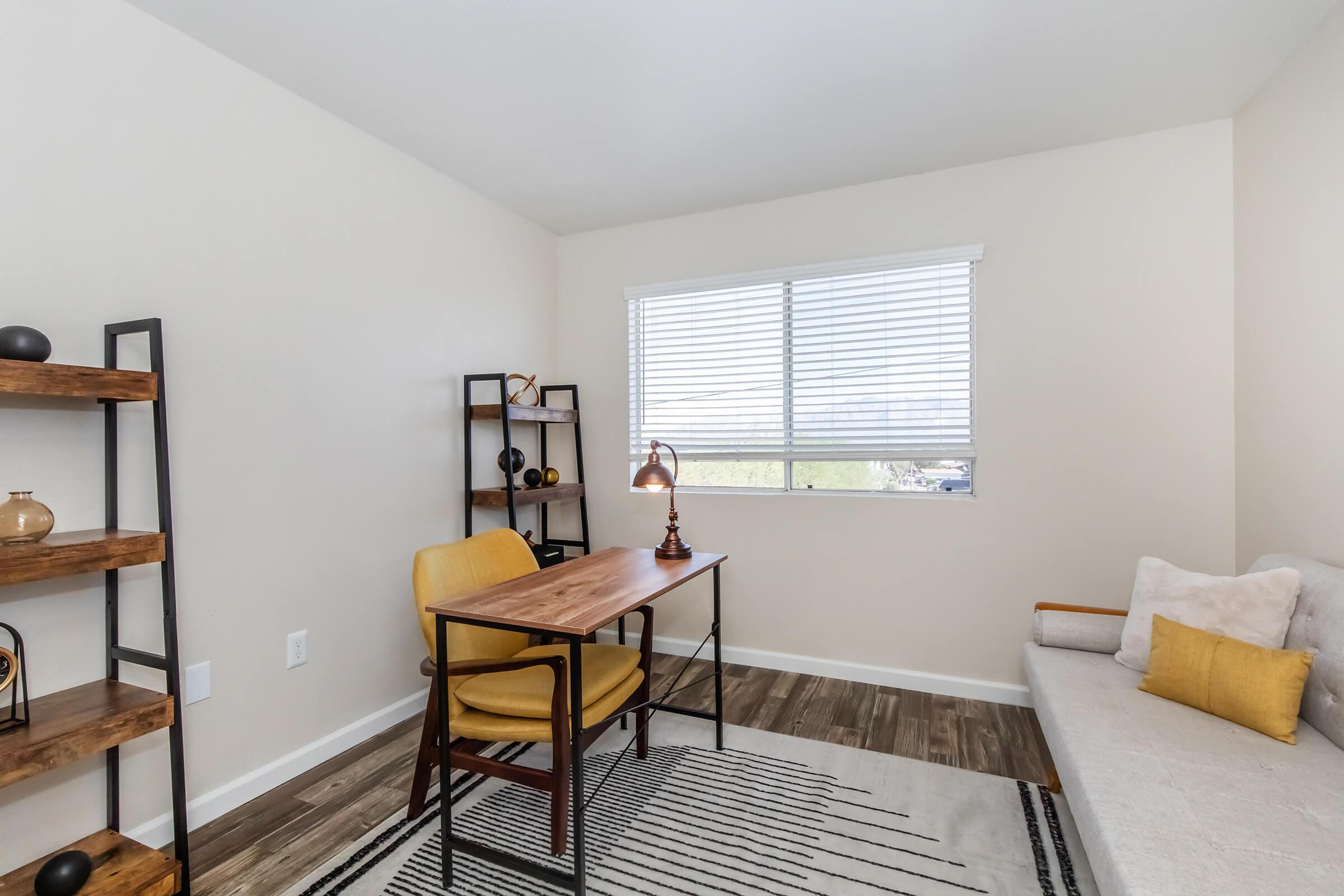 A cozy office space featuring a wooden desk with a stylish lamp, a yellow armchair, and a light-colored sofa. A tall bookshelf holds decorative items, and sunlight streams through a window with blinds, creating a warm and inviting atmosphere. The flooring is wooden, and a patterned rug adds texture to the room.