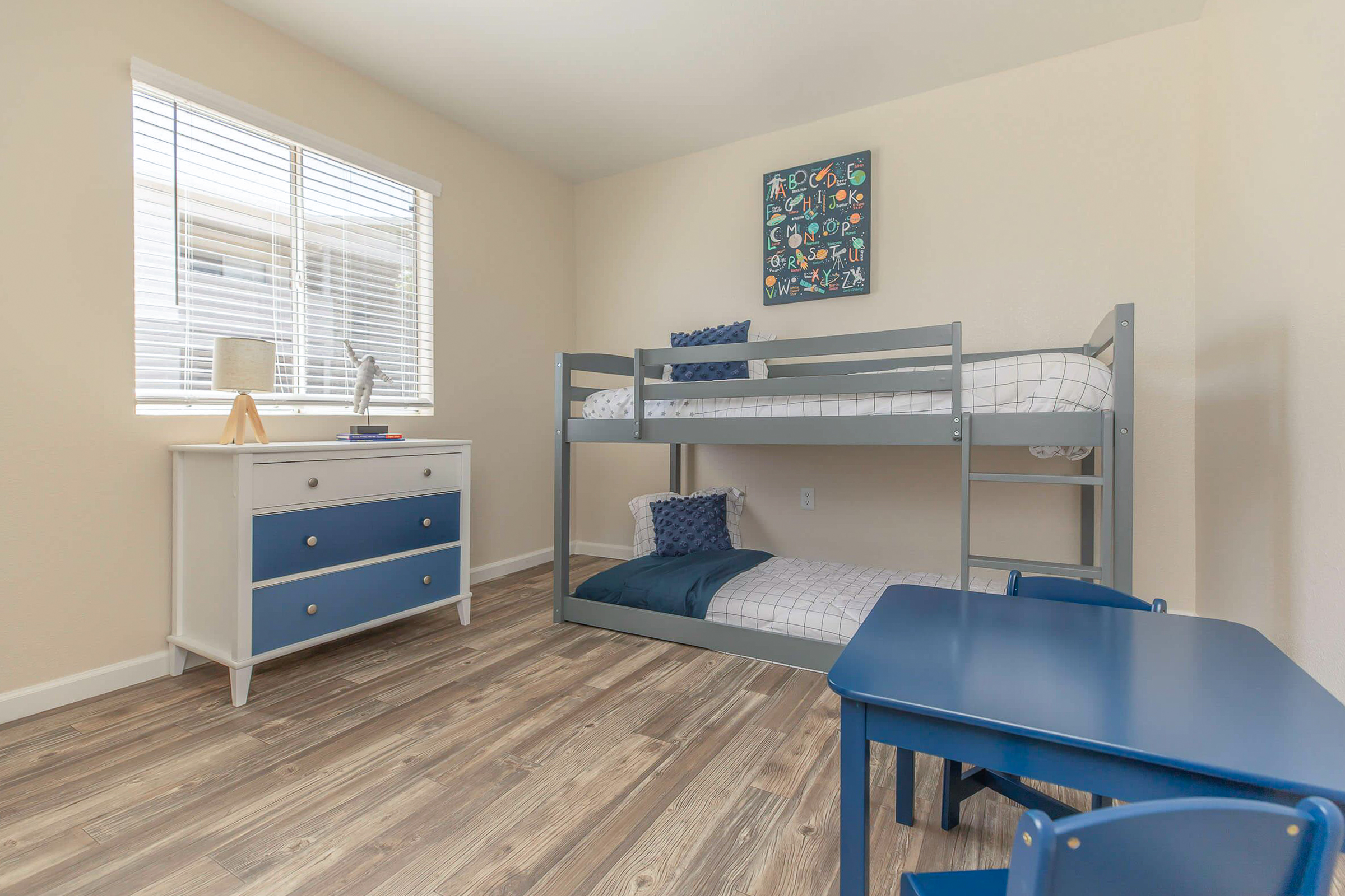 A brightly lit room featuring a gray bunk bed with a blue and white bedding set. A small blue table with two chairs sits on the wooden floor. A white dresser with blue handles is against the wall, and a decorative wall art piece is above the bunk bed. A window allows natural light to enter the space.