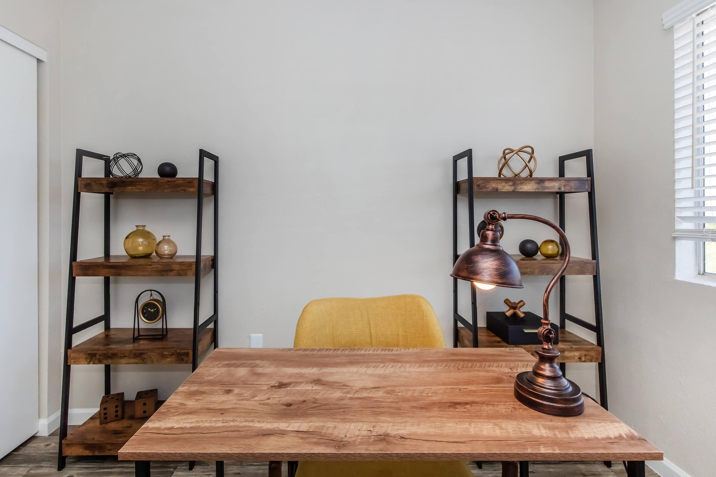 A modern workspace featuring a wooden desk with a bronze desk lamp, a yellow chair, and two black metal shelves on either side. The shelves display decorative items like gold and black spheres, a clock, and geometric shapes, against a light-colored wall with a window.