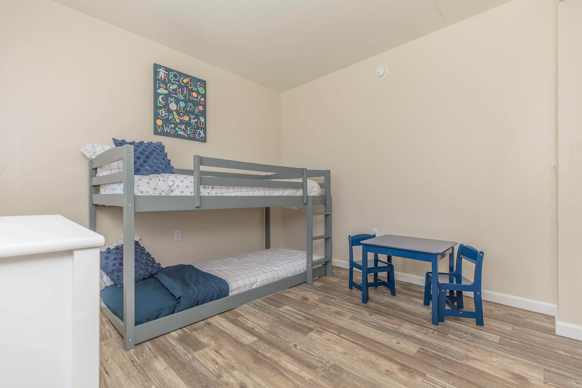A cozy children's room featuring a gray metal bunk bed with blue bedding, a small blue table with two matching chairs, and a colorful wall art piece. The room has light-colored walls and wooden flooring, creating a warm and inviting atmosphere.