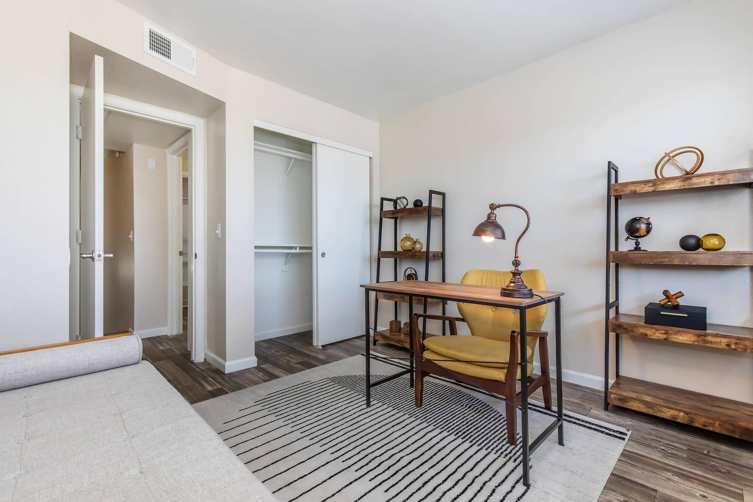 A modern workspace featuring a wooden desk and a yellow chair, with a desk lamp providing light. Behind the desk are shelves displaying decorative items. A closet is visible through an open door, and the room has a neutral color palette and wood-like flooring, creating a cozy atmosphere.