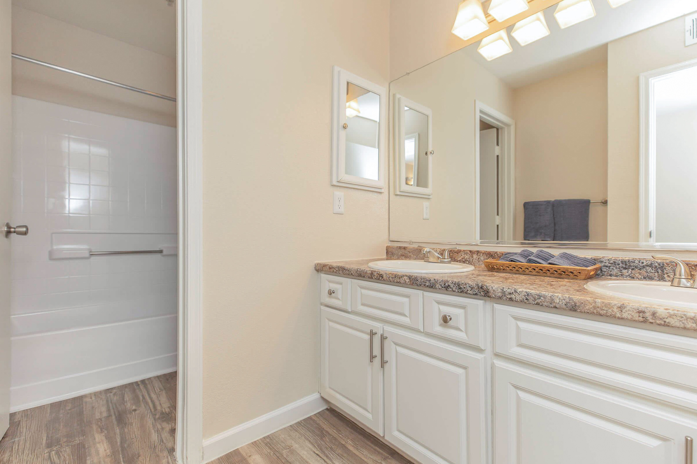 A modern bathroom featuring a double sink vanity with a granite countertop, two mirrors, and neatly arranged blue towels. The shower area is partially visible with white tiles. The walls are painted a light color, and the floor has a wood-like finish, creating a clean and inviting atmosphere.
