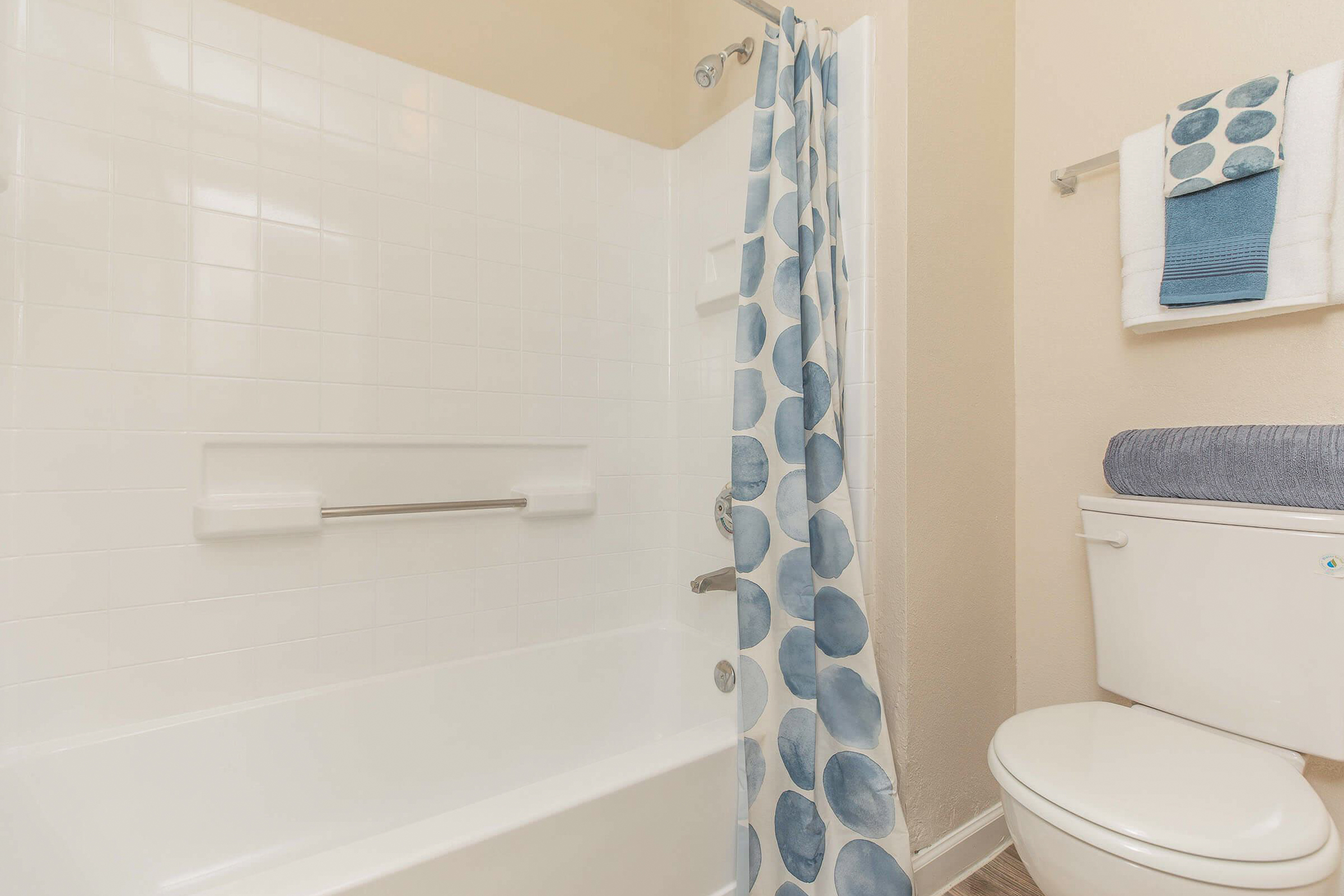 A clean bathroom featuring a bathtub with a shower, light-colored tiles, a blue and white patterned shower curtain, a towel rack with neatly folded towels, and a toilet. The walls are painted a warm beige.