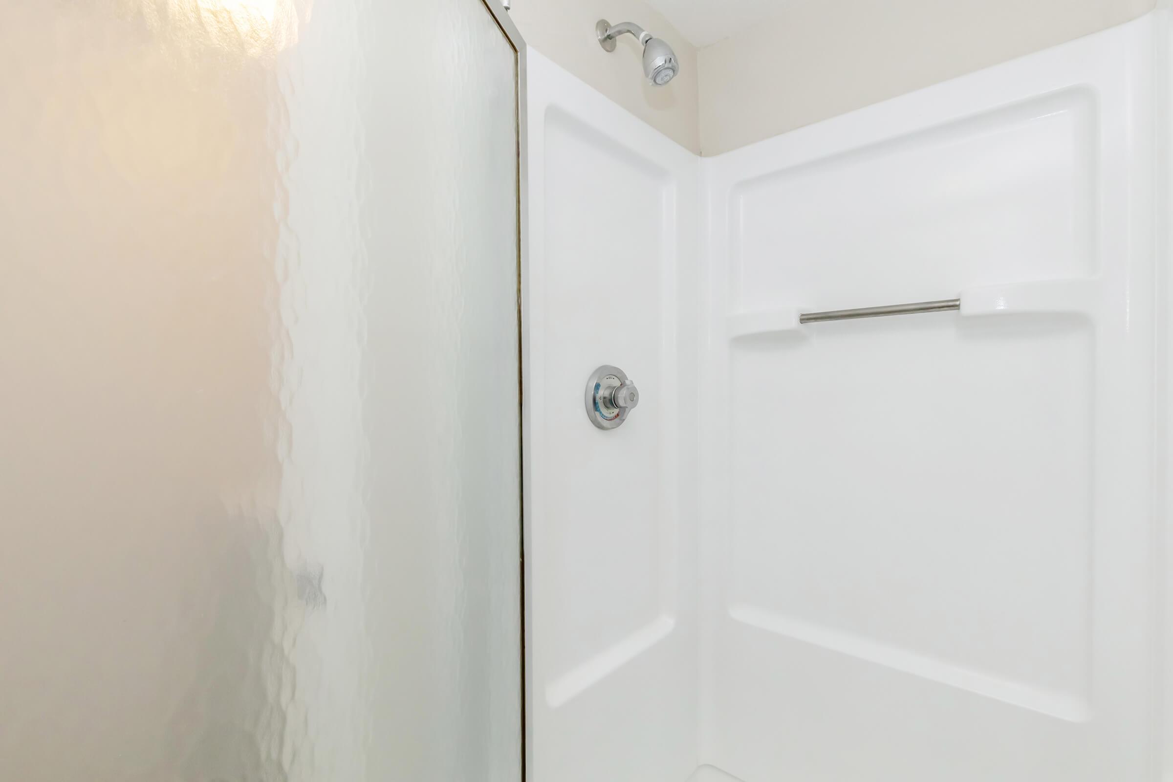 A clean and modern shower stall with a frosted glass door and a white acrylic interior. The shower features a chrome showerhead and a grab bar on the wall. The overall design is simple and functional, ideal for a bathroom setting.