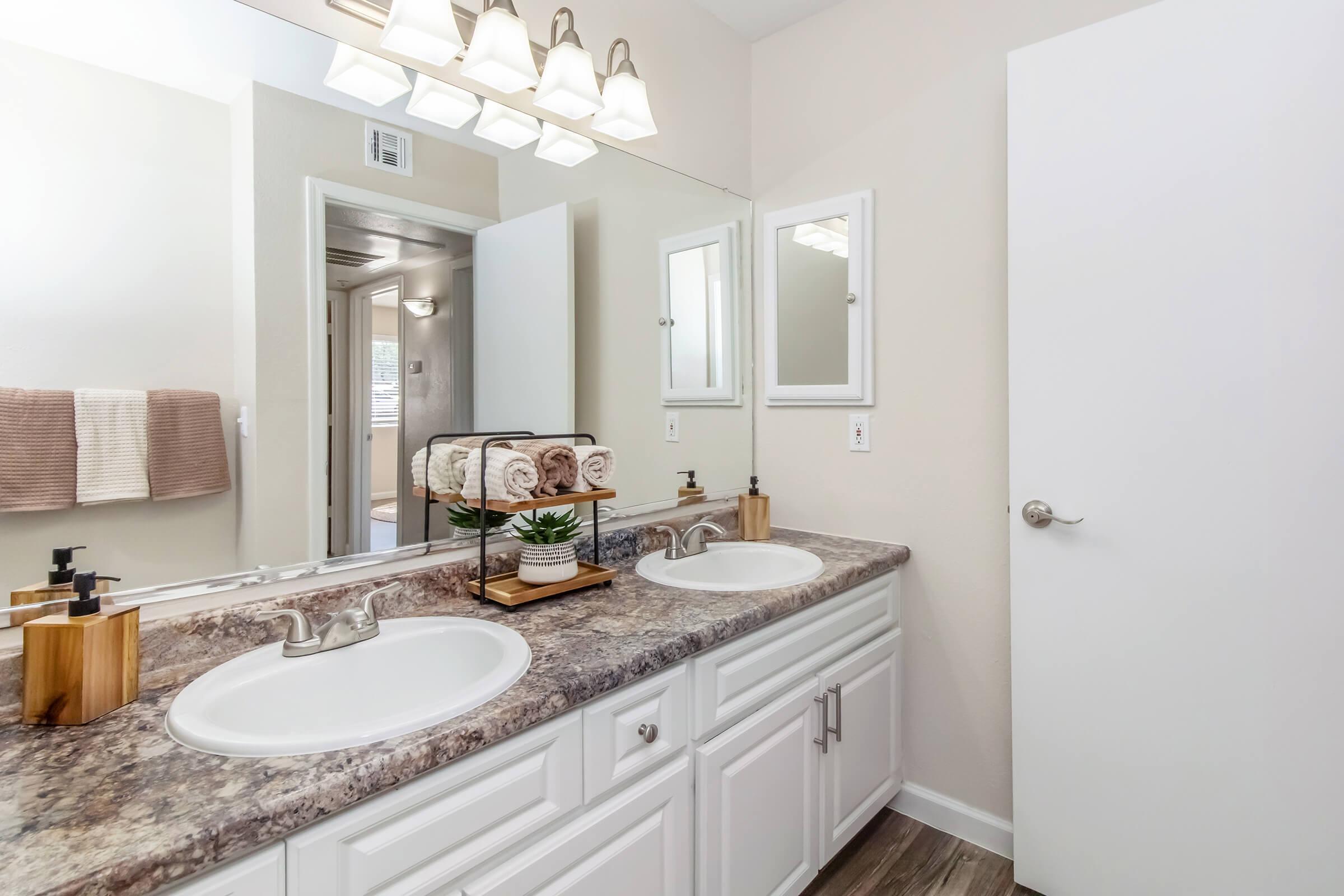A modern bathroom with a double sink vanity featuring two faucets, a stone countertop, and a decorative tray with rolled towels and a small plant. Soft lighting from above enhances the clean, neutral color scheme, and there are mirrors above each sink along with a door leading to another room.
