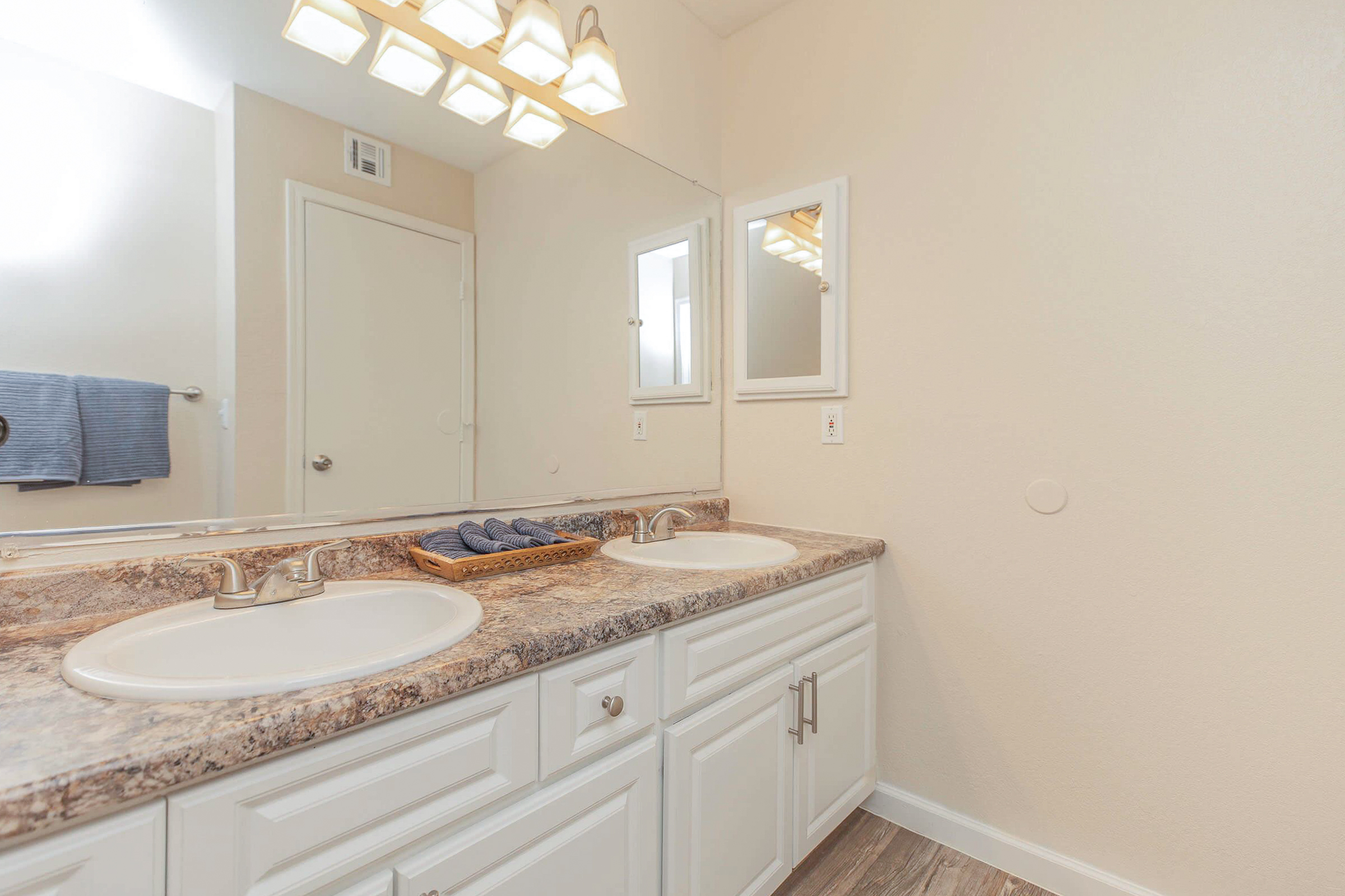 A bright, modern bathroom featuring dual sinks with polished faucets and a speckled countertop. Above the sinks, there are two mirrors reflecting the space, accompanied by warm overhead lighting. A towel rack with a blue towel and a basket of rolled towels add a decorative touch. The walls are a soft neutral color.