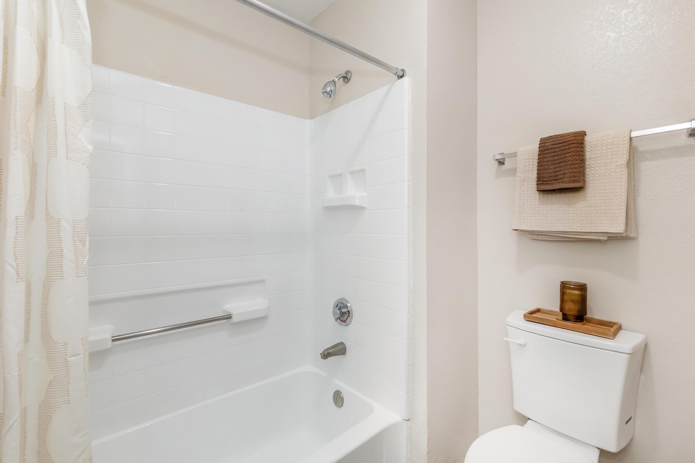 A clean and minimalistic bathroom featuring a white bathtub with shower, a light-colored shower curtain, and neatly arranged towels on a towel rack. A small wooden tray with a candle is placed on the toilet tank, adding a touch of warmth to the space. The walls are painted in a soft, neutral tone.