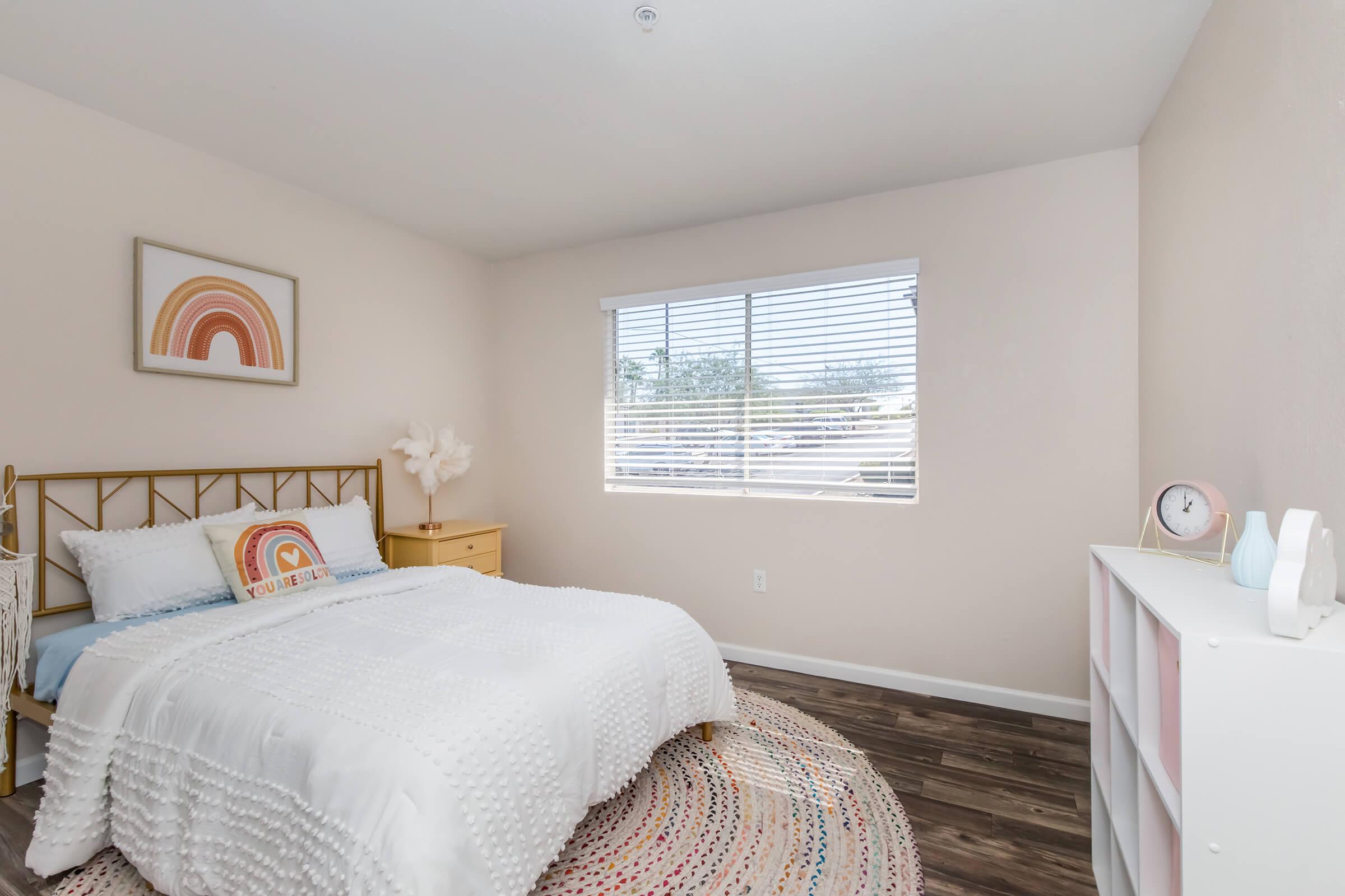 A cozy, well-lit bedroom featuring a gold-framed bed with white bedding, a decorative rainbow print on the wall, a nightstand, and a round rug. Natural light streams through a window with shades. The flooring is dark wood, and a white bookshelf is positioned in the corner.