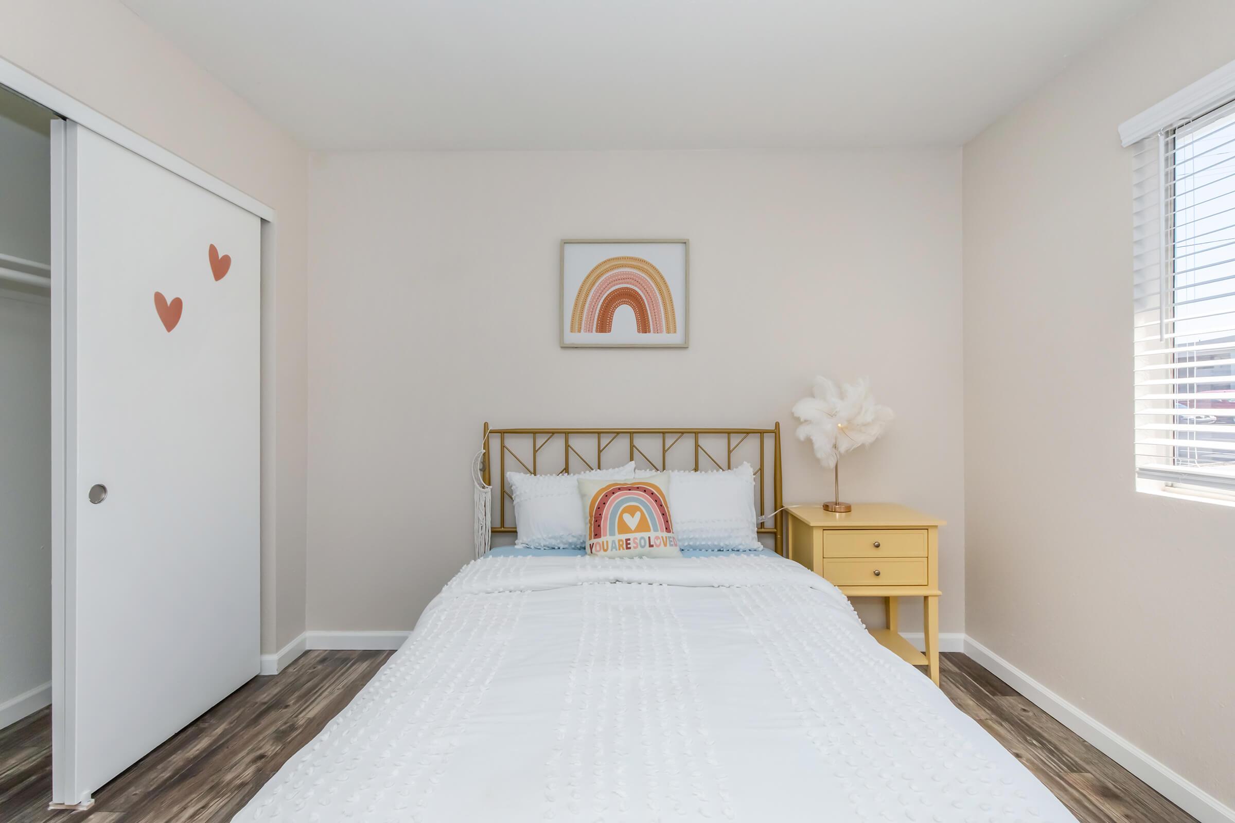 A cozy bedroom featuring a white bed with a decorative rainbow pillow, a yellow nightstand with a lamp, and a wall adorned with heart-shaped decorations. There's a closet door on the left and a window with blinds, allowing natural light to brighten the room. The flooring is a warm wood finish.