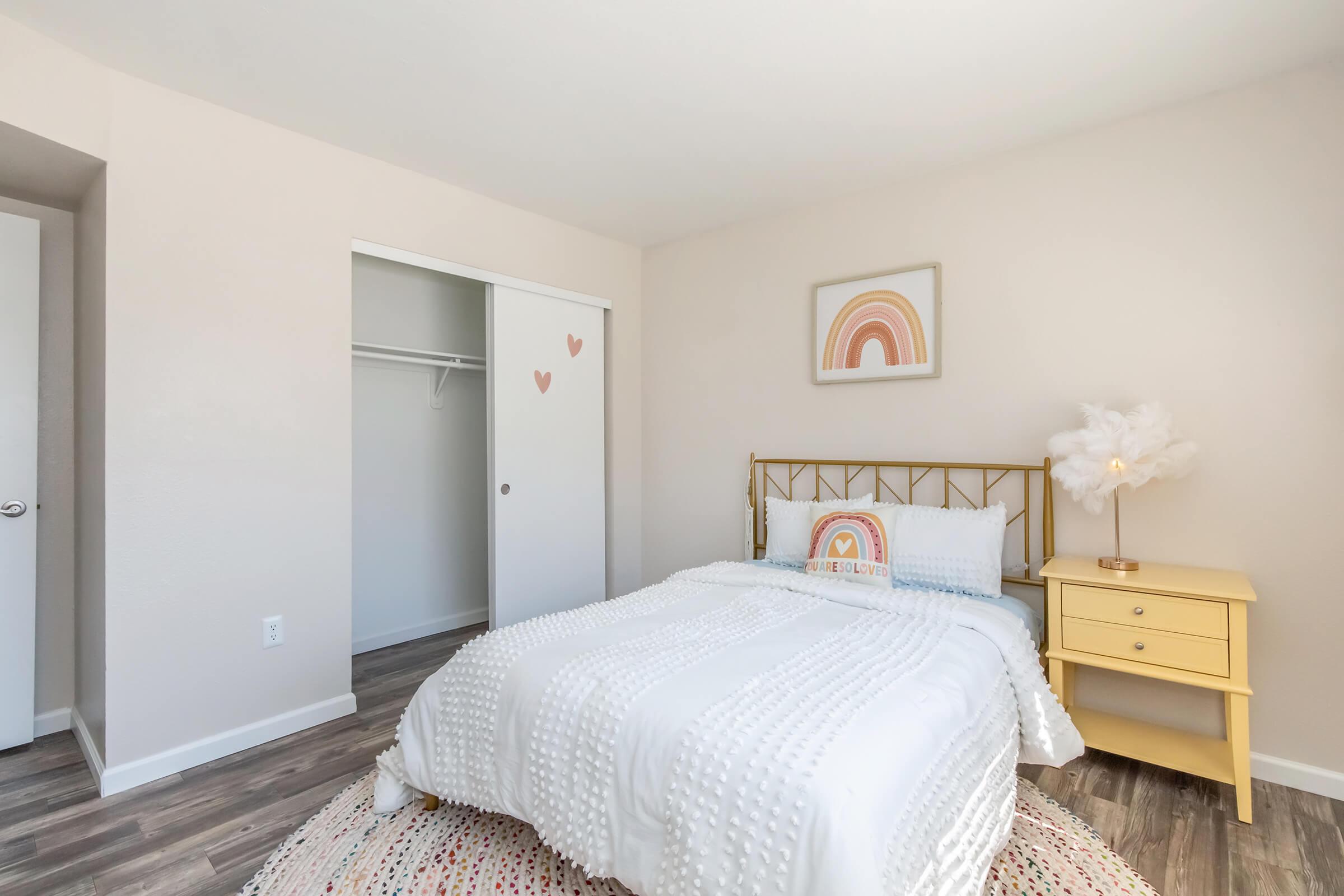 A cozy, well-lit bedroom featuring a bed with a white textured duvet, a yellow nightstand with a lamp, and a decorative rainbow artwork on the wall. The room has a light beige color scheme and hardwood flooring, along with a closet with sliding doors. Heart-shaped decor adds a playful touch.