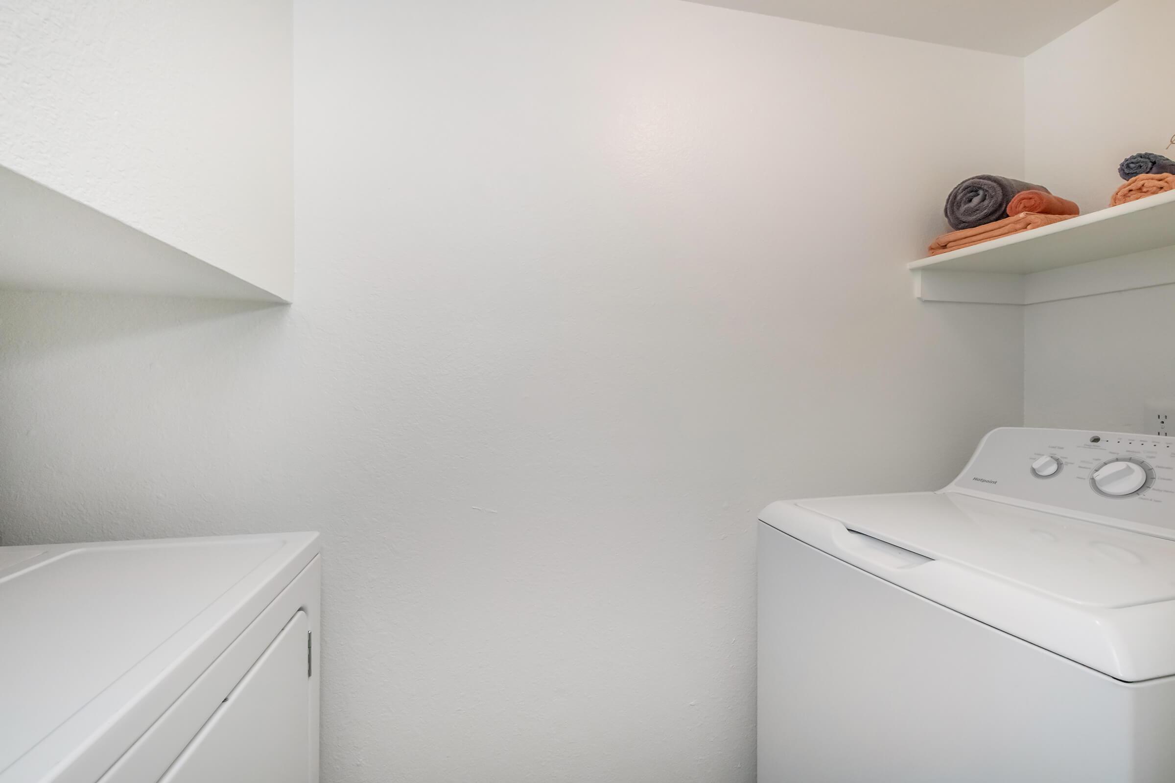 A clean laundry area featuring a white washing machine and dryer. Above the appliances, there are neatly stacked towels in various colors on a shelf. The walls are plain white, creating a bright and airy atmosphere in the space.