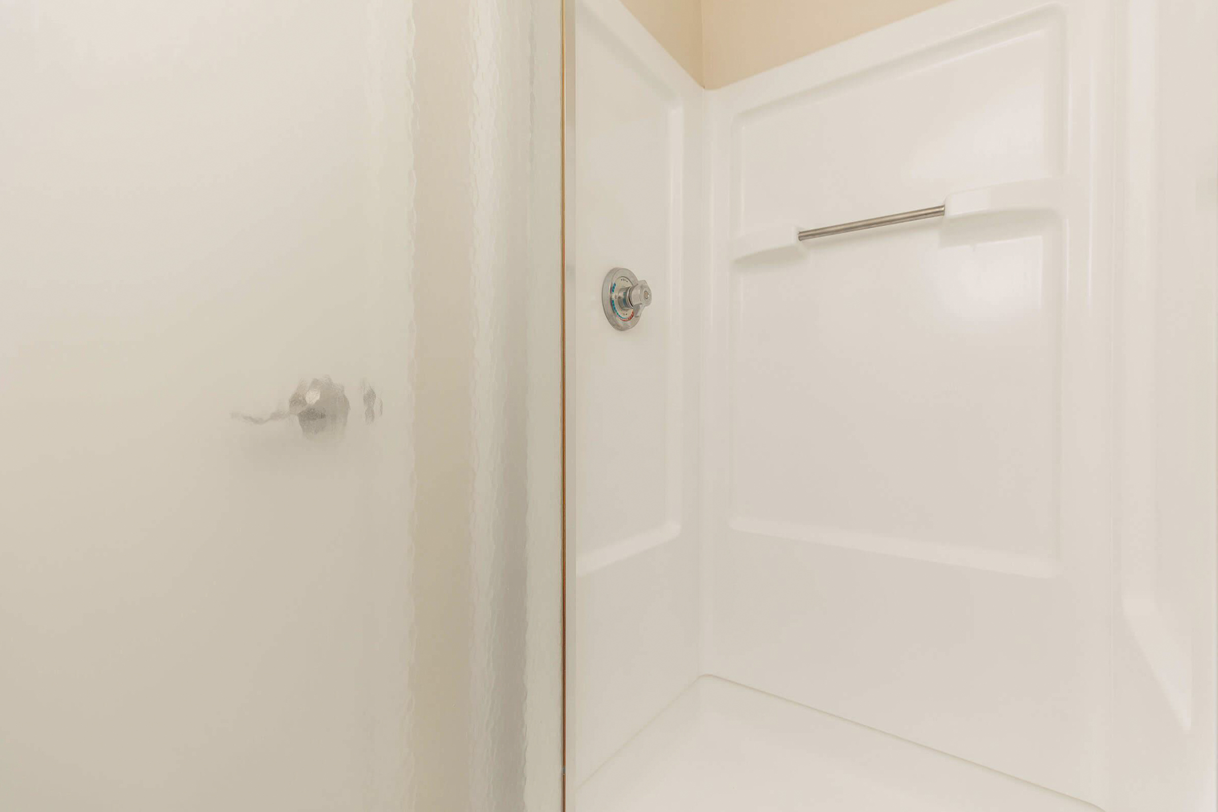 A modern shower stall featuring a textured glass door and white, tiled walls. The shower includes a chrome valve and a grab bar for stability. The overall design emphasizes cleanliness and simplicity in a bright, minimalistic space.