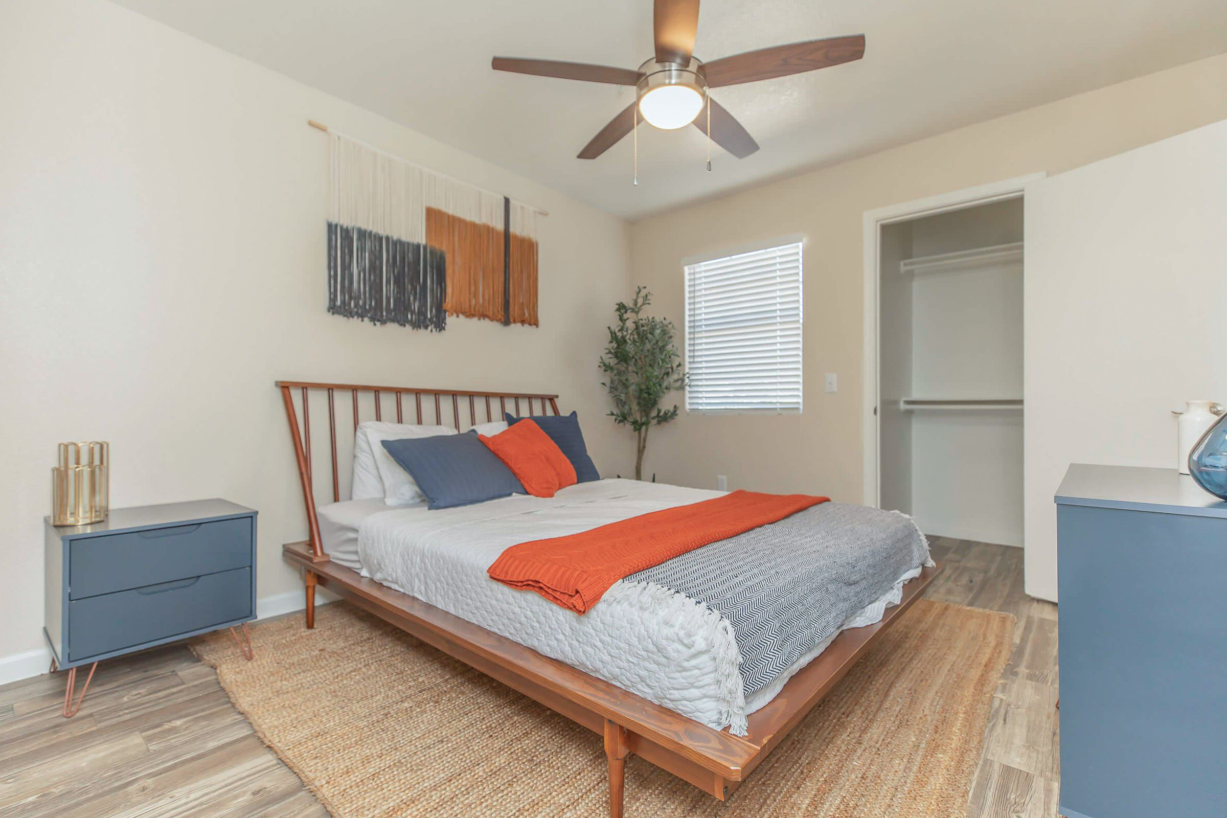 A cozy bedroom featuring a wooden bed frame with a gray and white bedding set, accented by an orange throw pillow. There are stylish nightstands on either side of the bed, a geometric area rug, and a wall hanging above the bed. A ceiling fan and a small potted plant add to the decor, with a closet visible in the background.