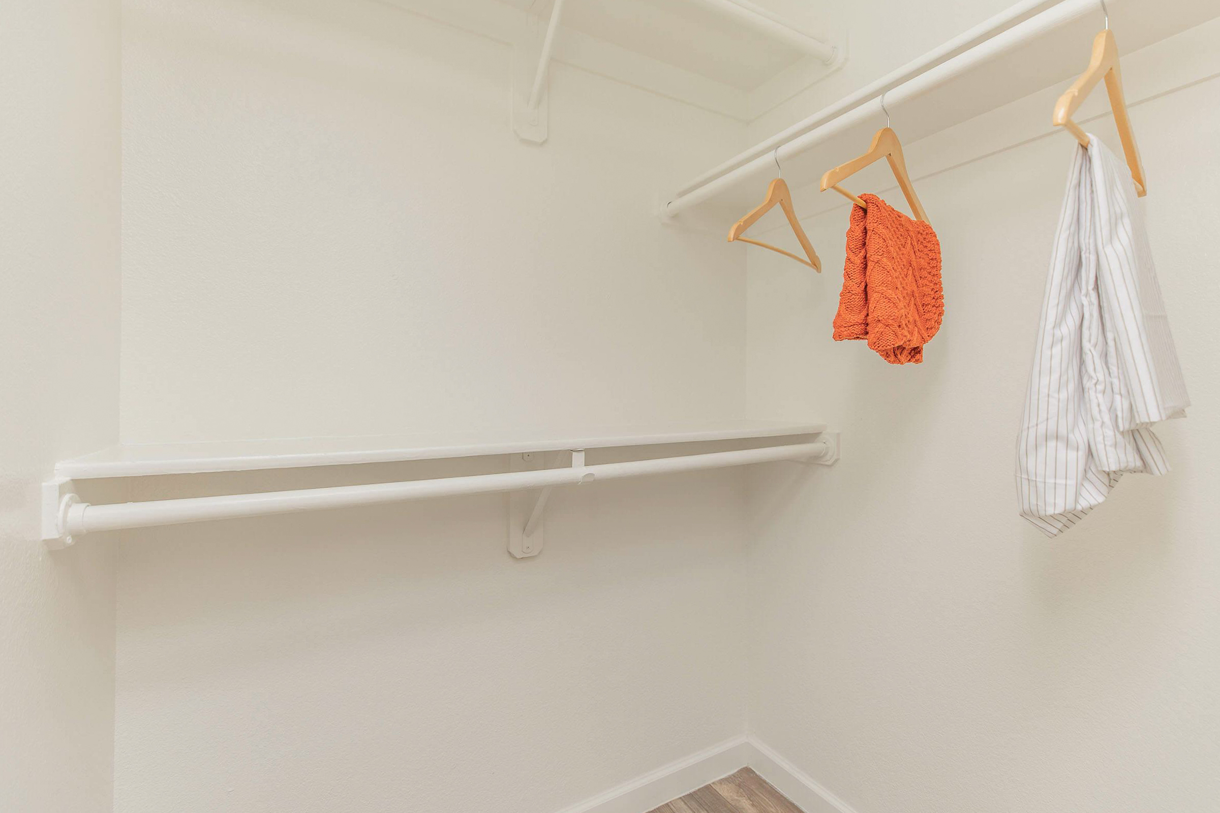 Empty closet with white walls and wooden flooring. A hanging rod with two wooden hangers holds a wrinkled white shirt and a bright orange piece of clothing. There is an unused space on the rod, indicating storage potential.