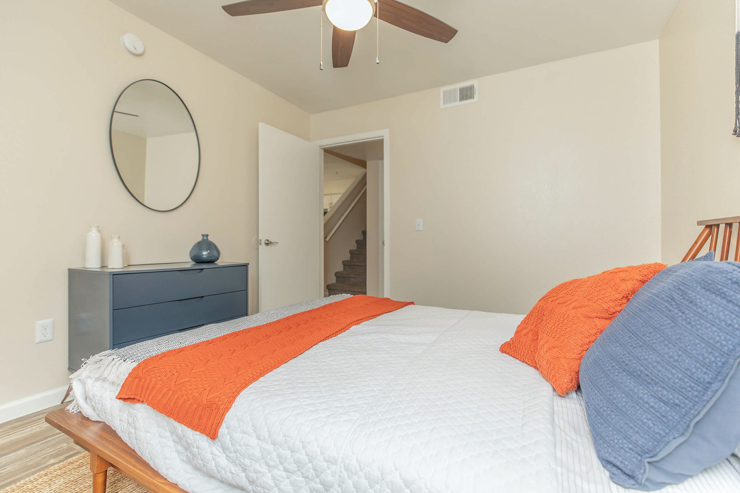 A cozy bedroom featuring a neatly made bed with an orange and blue accent pillow, a modern dresser, a round mirror on the wall, and a ceiling fan. The room has light-colored walls and an open door that leads to a staircase, creating an inviting ambiance.