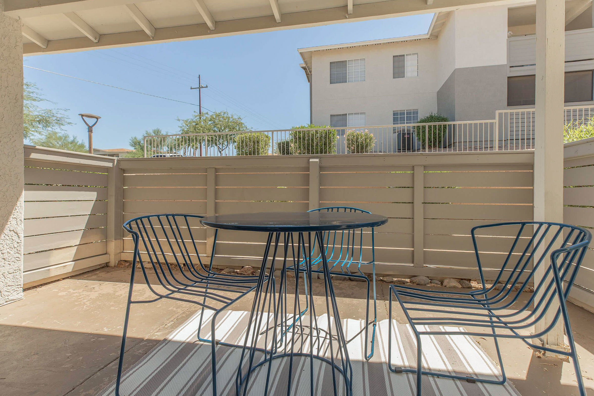 A patio area featuring a round black table surrounded by four blue wire chairs, set on a striped rug. In the background, there is a gray building with windows and a fence. The scene is illuminated by sunlight, with clear skies and greenery visible beyond the fence.