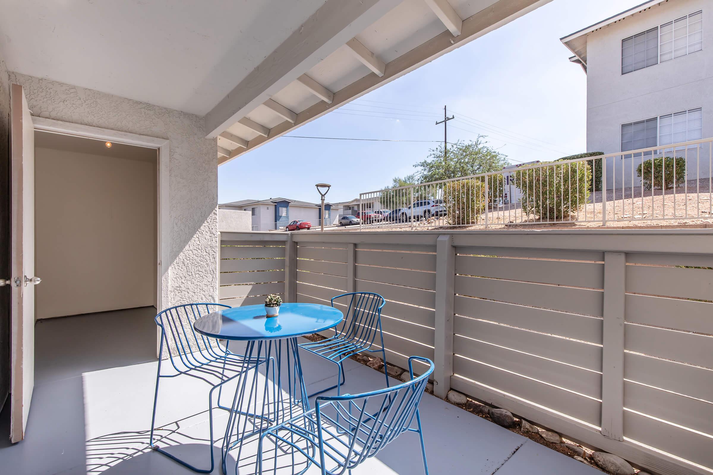 A small outdoor patio area featuring a blue metal table and matching chairs, with a potted plant on the table. The patio is enclosed by a wooden fence, and in the background, there are parked cars and building structures under a clear blue sky.