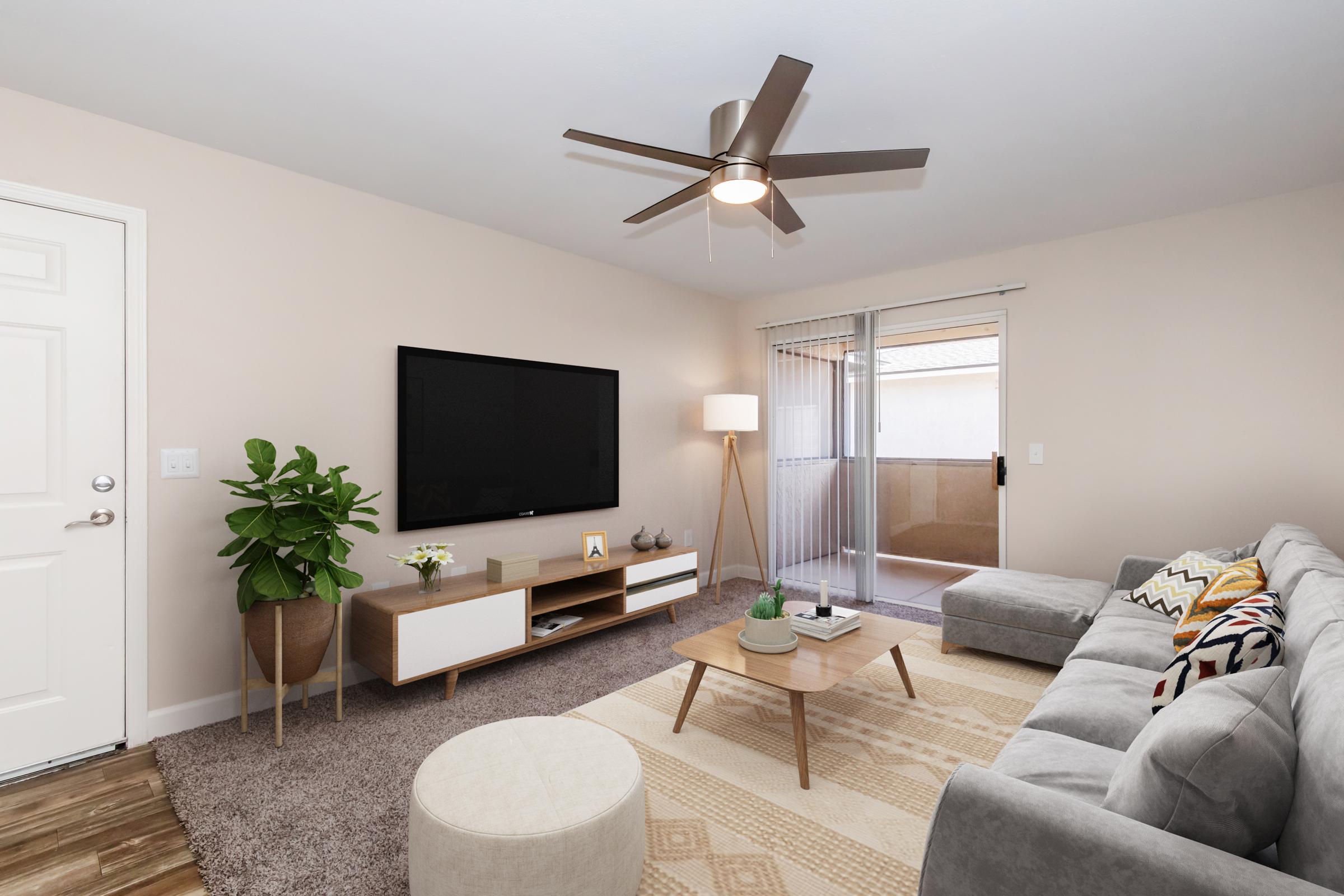 A cozy living room featuring a gray sectional sofa with decorative cushions, a coffee table, and a light-colored rug. There's a TV mounted on the wall above a modern console cabinet. A potted plant sits beside the sofa, and a patio door leads to an outdoor space. A ceiling fan provides cooling.