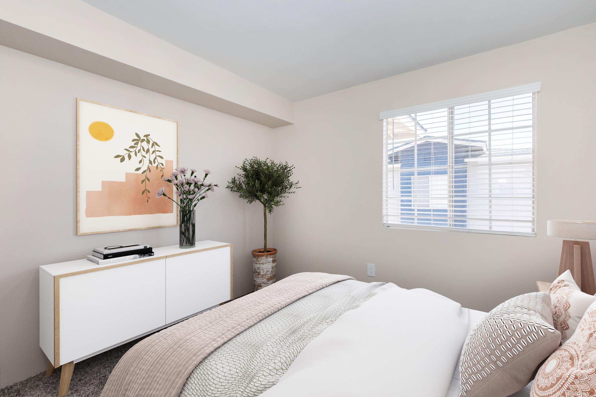 A bright, modern bedroom featuring a neatly made bed with light bedding, a decorative pillow, and a bedside lamp. There's a white dresser against the wall, adorned with a vase of flowers, and an abstract art piece hanging above it. Natural light streams in through a window with white blinds, showcasing a view outside.