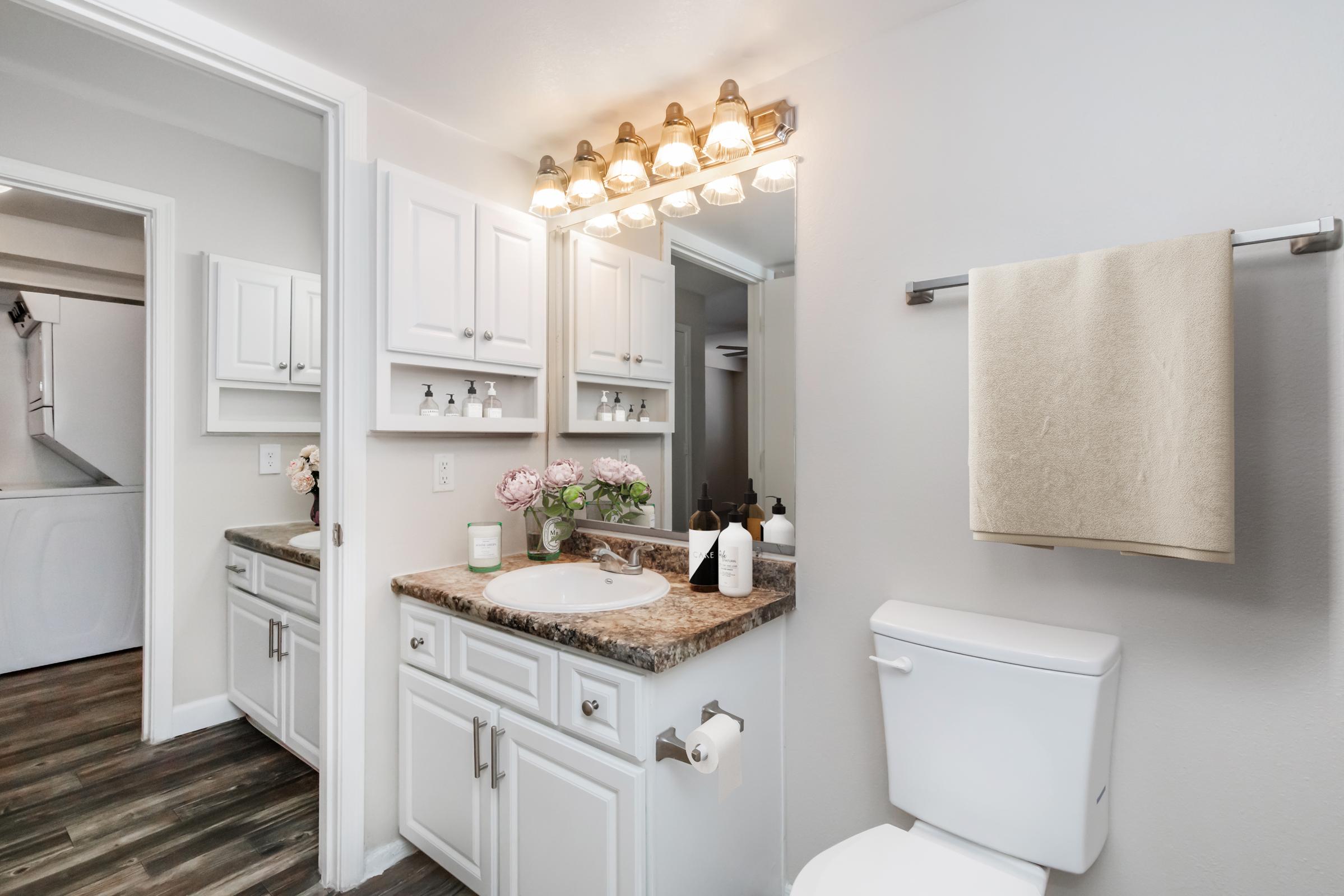 A modern bathroom featuring a double vanity with a granite countertop, two sinks, and ample cabinet storage. A large mirror with stylish lighting above. To the right, there's a toilet and a towel rack holding a beige towel. Fresh flowers add a decorative touch, and the flooring is a wood-look design.