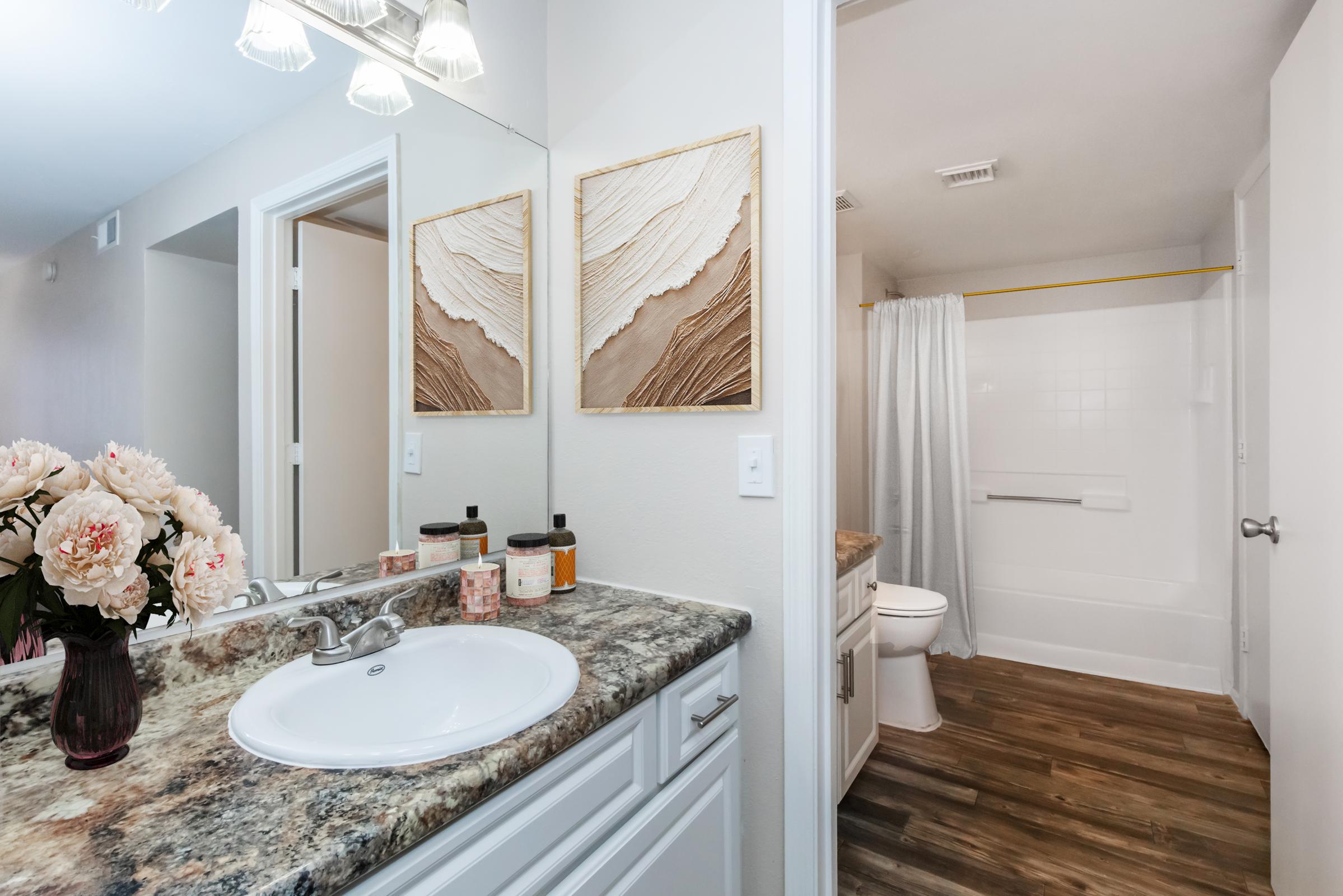 A well-lit bathroom featuring a double sink vanity with a granite countertop, decorative wall art, and a mirror. The tiled floor complements the clean aesthetic. A shower area with a white curtain is visible in the background, adding to the modern design of the space. Fresh flowers add a touch of elegance.