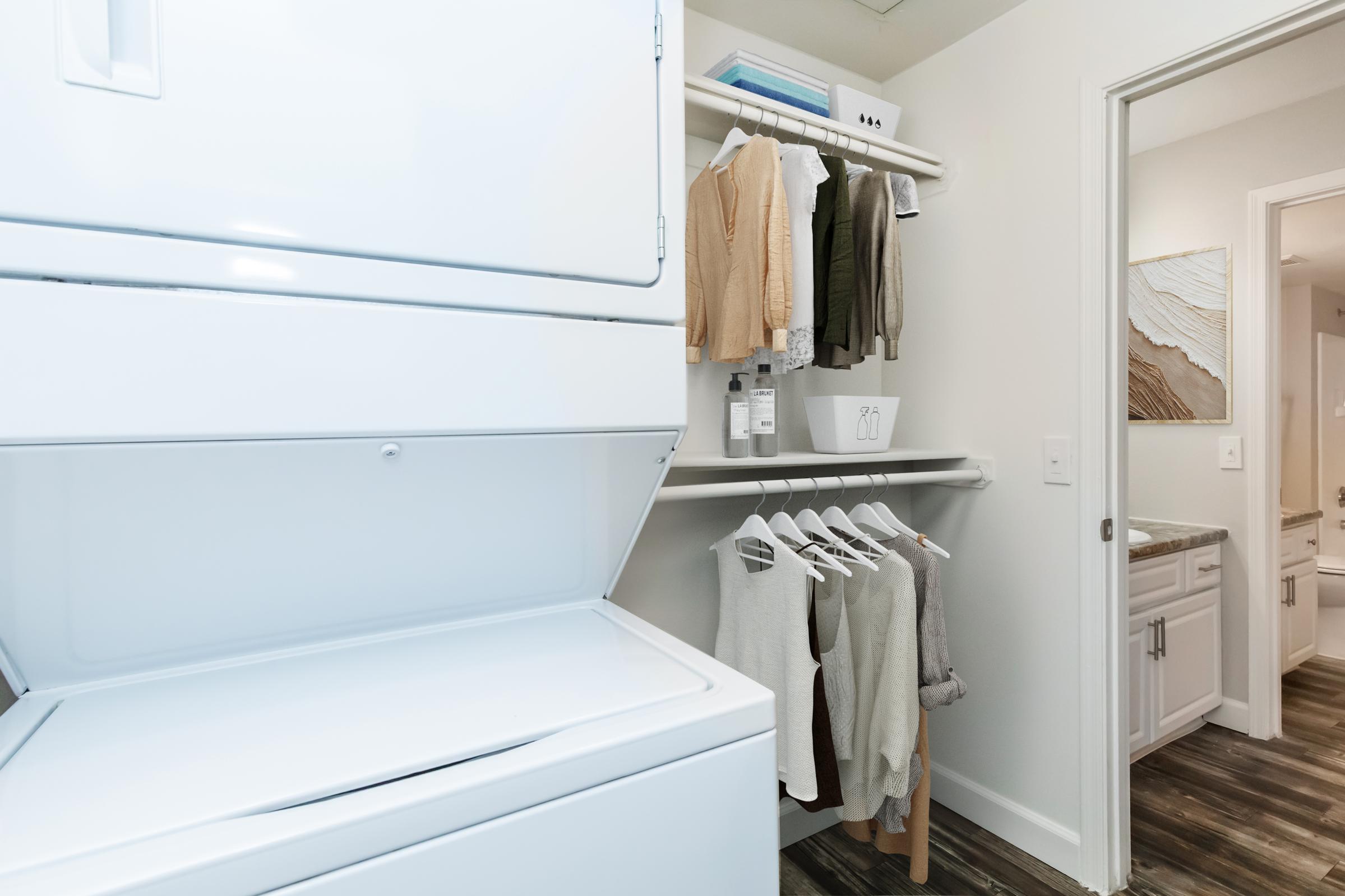 A modern laundry room featuring a stacked washer and dryer on the left. To the right, there is a shelf with neatly folded items and a rod holding several hanging clothes. In the background, a doorway leads to a bathroom with light-colored walls and cabinetry.