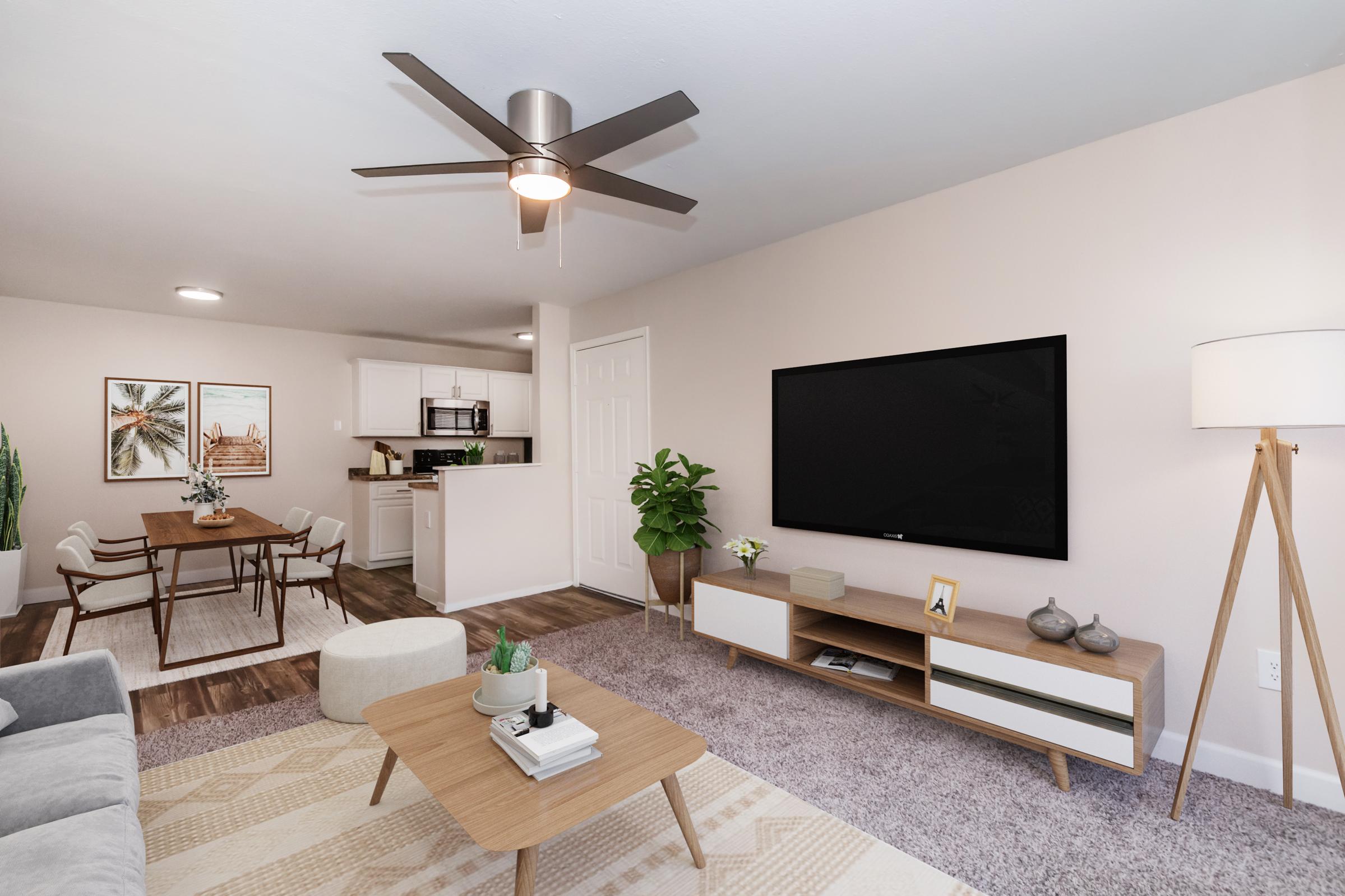 A modern living room featuring a light gray sofa, a coffee table, and a large TV mounted on the wall. There's a decorative plant next to the TV and a floor lamp. In the background, a dining area with a table and chairs is visible, along with a kitchen space. The decor is minimalistic and contemporary.