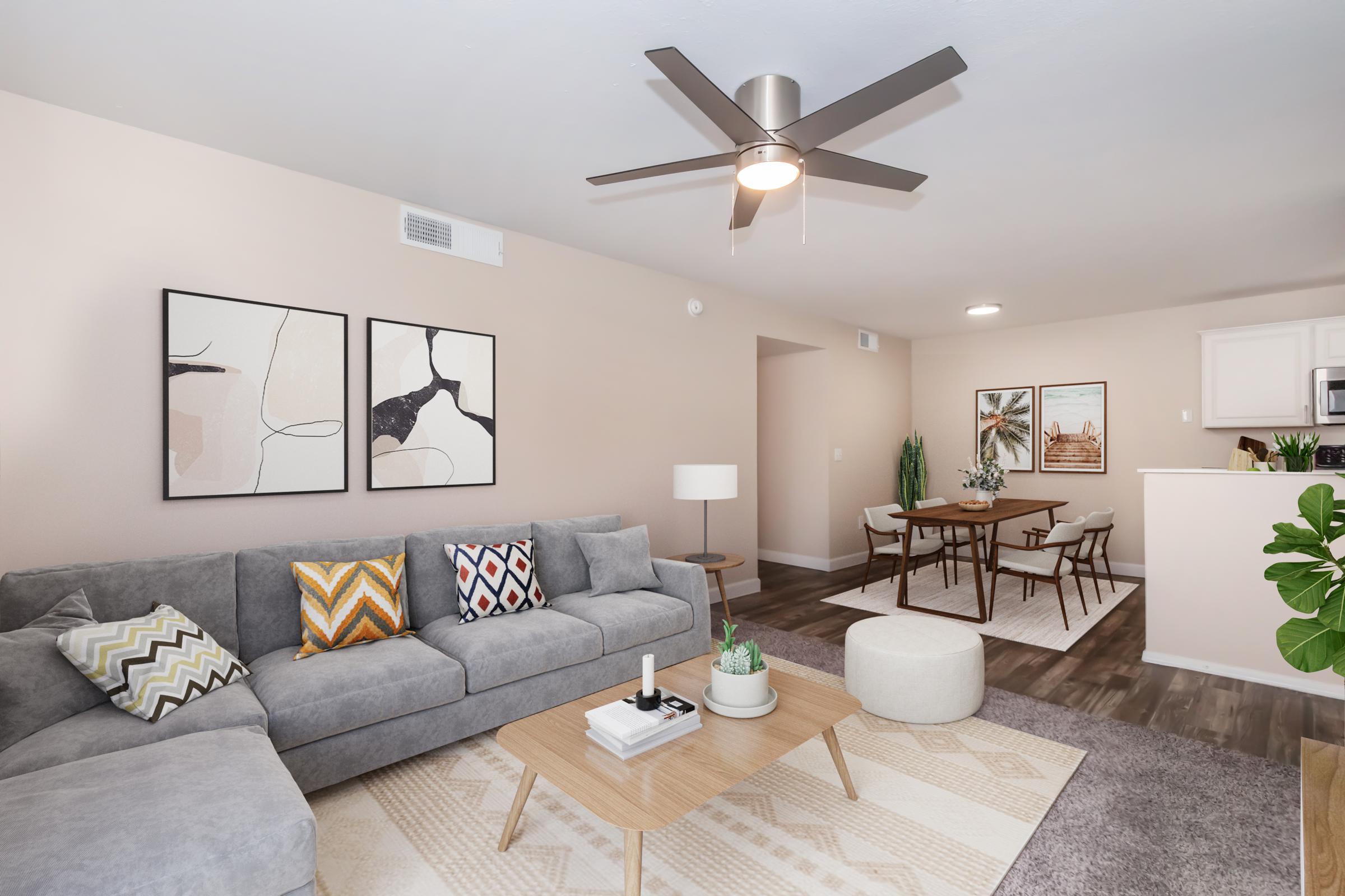 A stylish living room featuring a gray sectional sofa with colorful throw pillows, a round coffee table, and a ceiling fan. The walls are painted a soft beige, and there's wall art above the sofa. In the background, there are plants and a dining area with a small table and chairs, creating a cozy atmosphere.