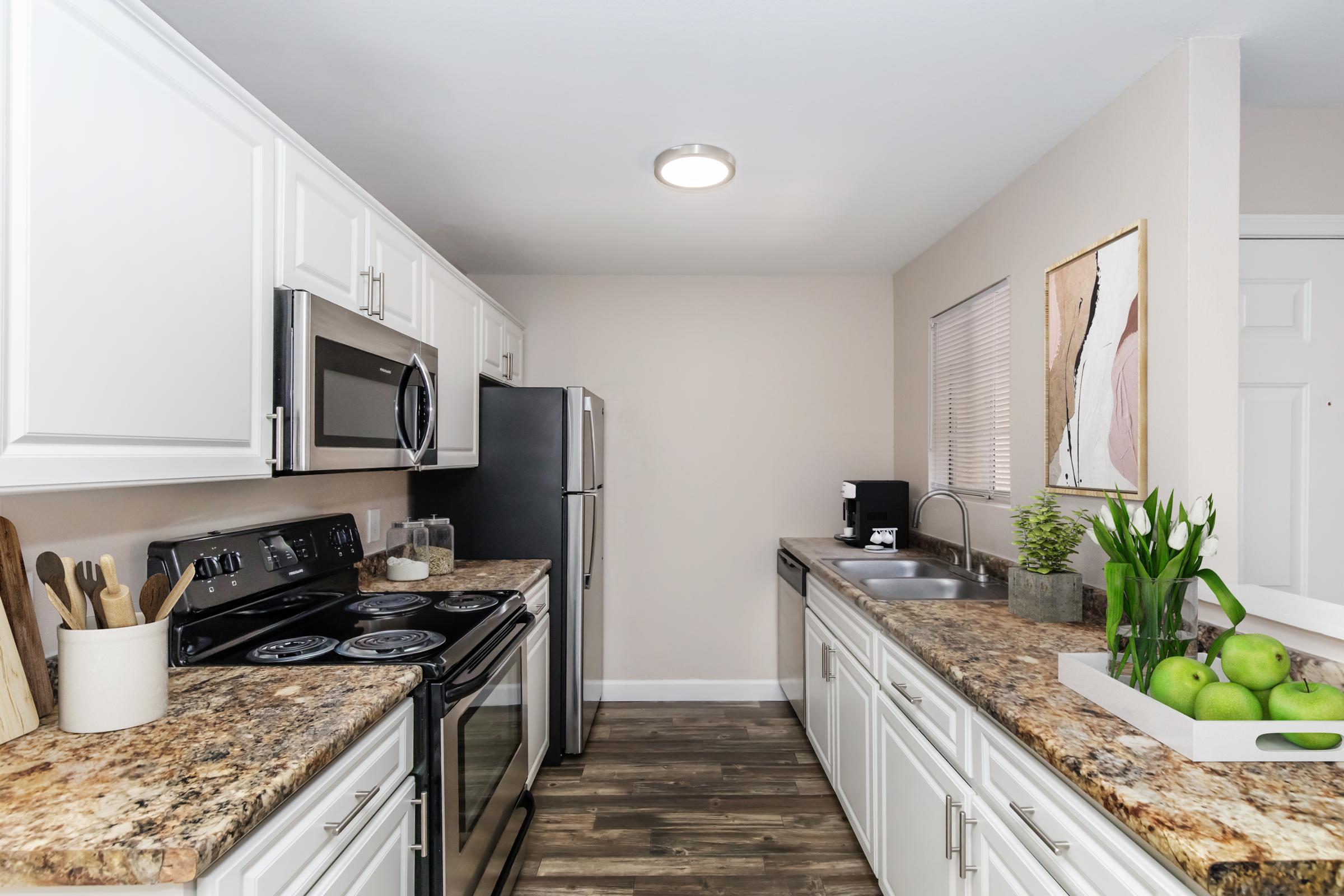 Modern kitchen featuring white cabinets, stainless steel appliances, and a granite countertop. Includes an electric stove, microwave, refrigerator, and a double sink. Decorative elements like kitchen utensils, a plant, and a bowl of green apples are arranged on the countertop, with neutral walls and natural lighting.