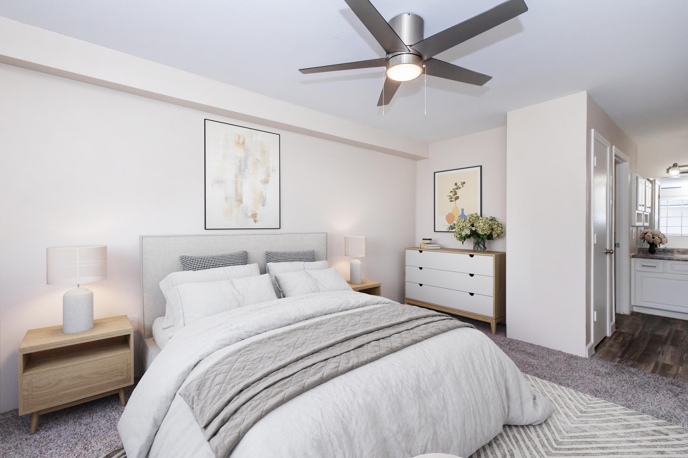A modern bedroom featuring a light-colored bed with gray and white bedding, two bedside lamps on wooden tables, a white dresser with decorative items, and soft carpeting. A ceiling fan provides ventilation. The walls are adorned with minimalist artwork and the room has a warm, inviting atmosphere.