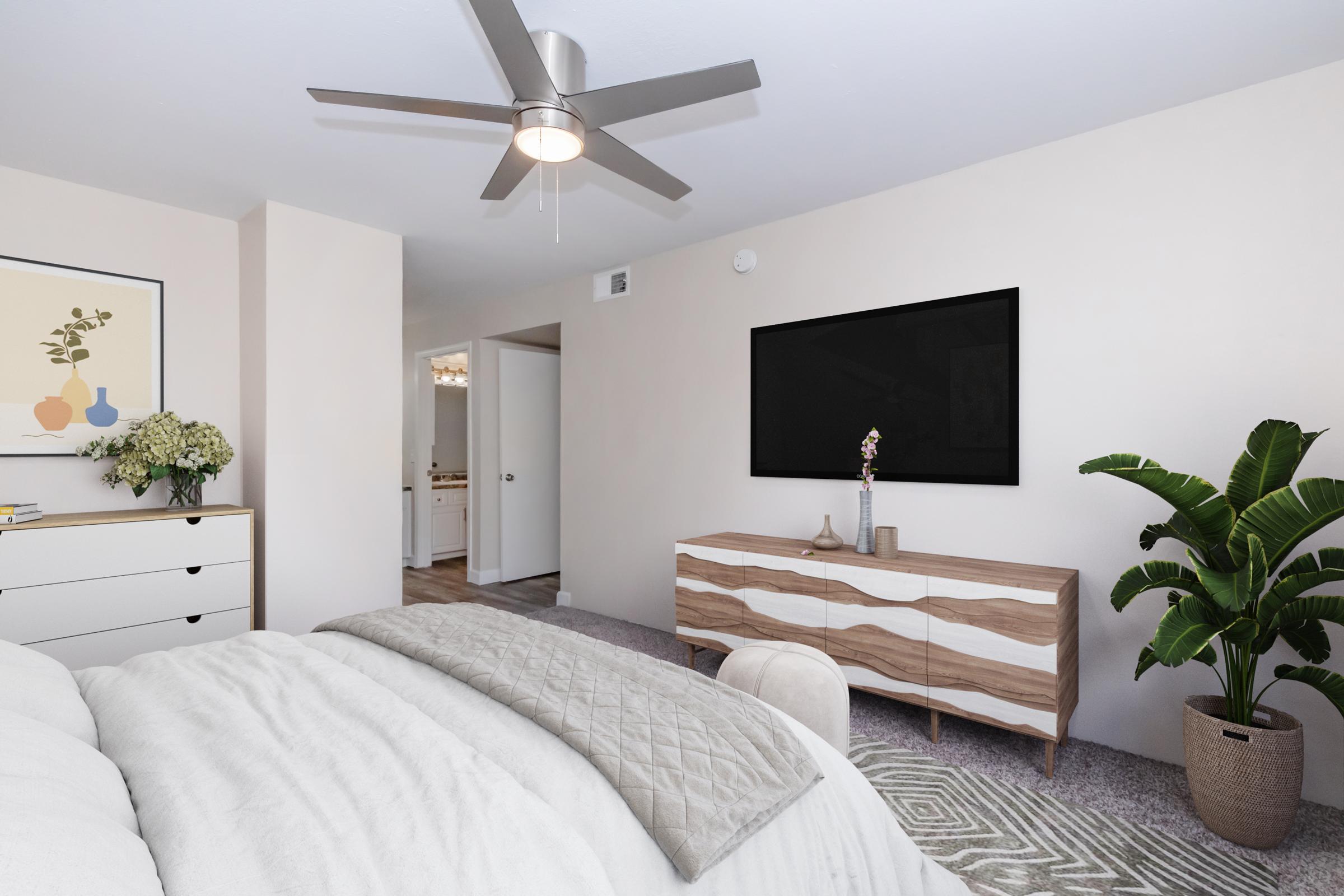 A modern bedroom featuring a light-colored wall, a large bed with a cozy blanket, a dresser with decorative items, a flat-screen TV mounted on the wall, and a green potted plant. A ceiling fan is visible, and a doorway leads to a bathroom in the background.