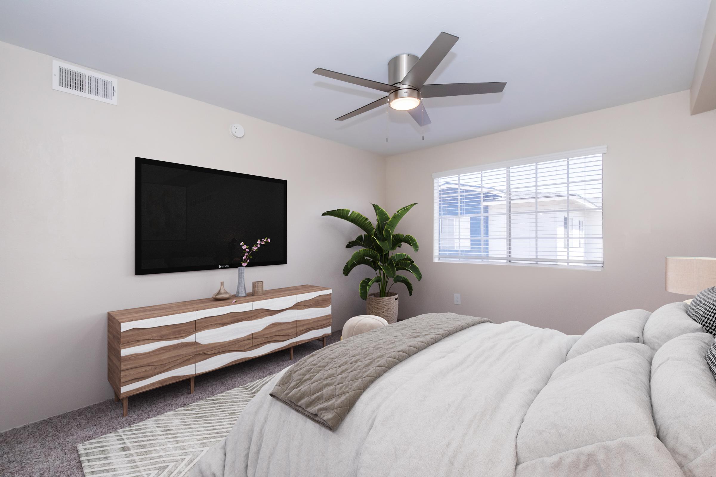 A cozy bedroom featuring a large window with white blinds, a ceiling fan, and a modern TV mounted on the wall. A stylish wooden dresser with a wavy design holds decorative items, while a green potted plant adds a touch of nature. The bedding is soft and inviting, contributing to a warm atmosphere.