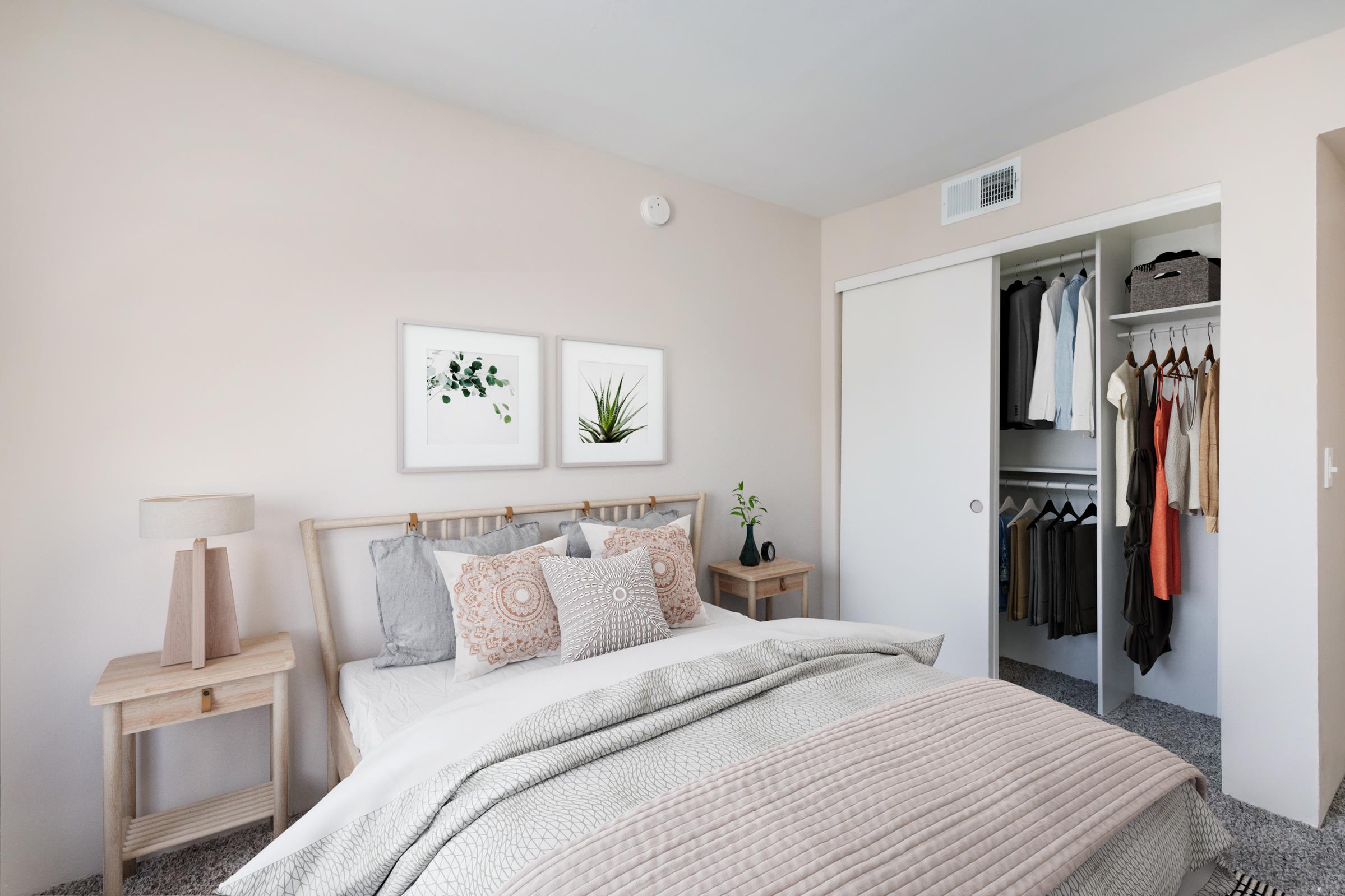 A cozy bedroom featuring a light color palette. The bed is adorned with decorative pillows and a textured blanket. Two framed botanical prints hang on the wall. A bedside table with a lamp stands next to the bed. In the closet, neatly hung clothes are visible, adding to the organized and inviting atmosphere.