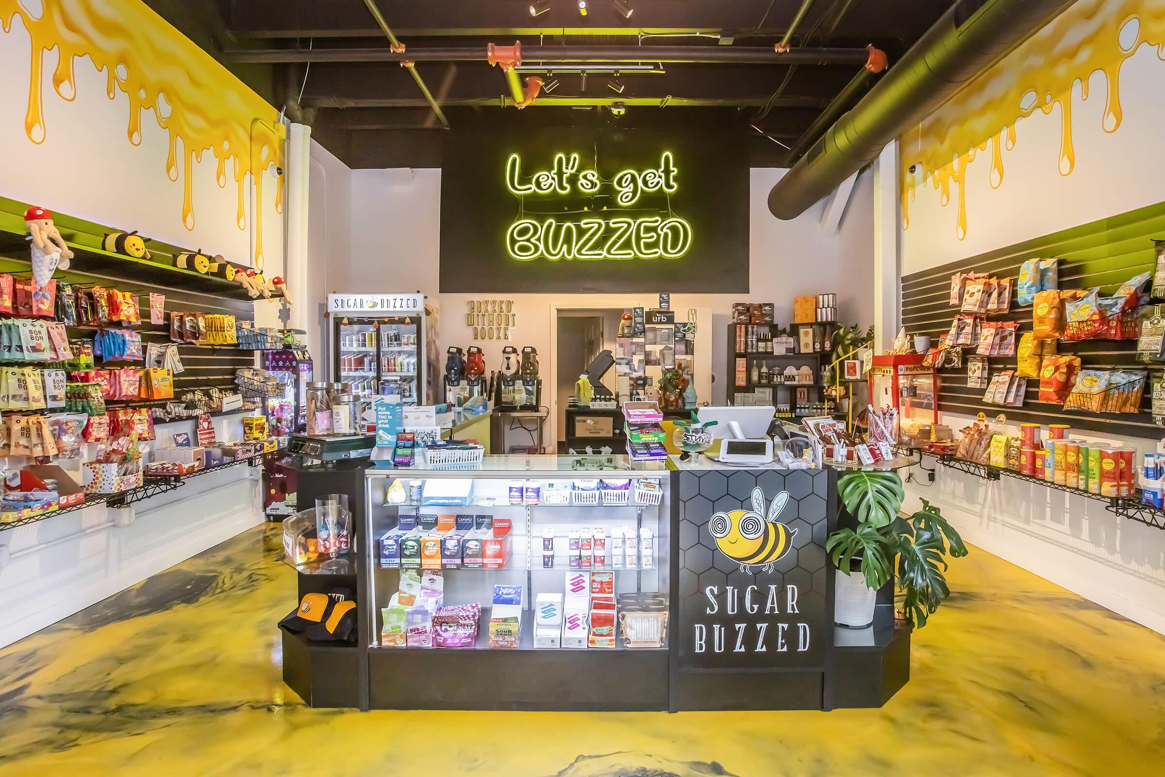 A brightly lit candy store with colorful shelves filled with snacks and sweets. The checkout counter at the front features various candies and beverages. A large neon sign above reads "Let's get BUZZED," and the floor has a playful yellow design, enhancing the cheerful atmosphere of the shop.