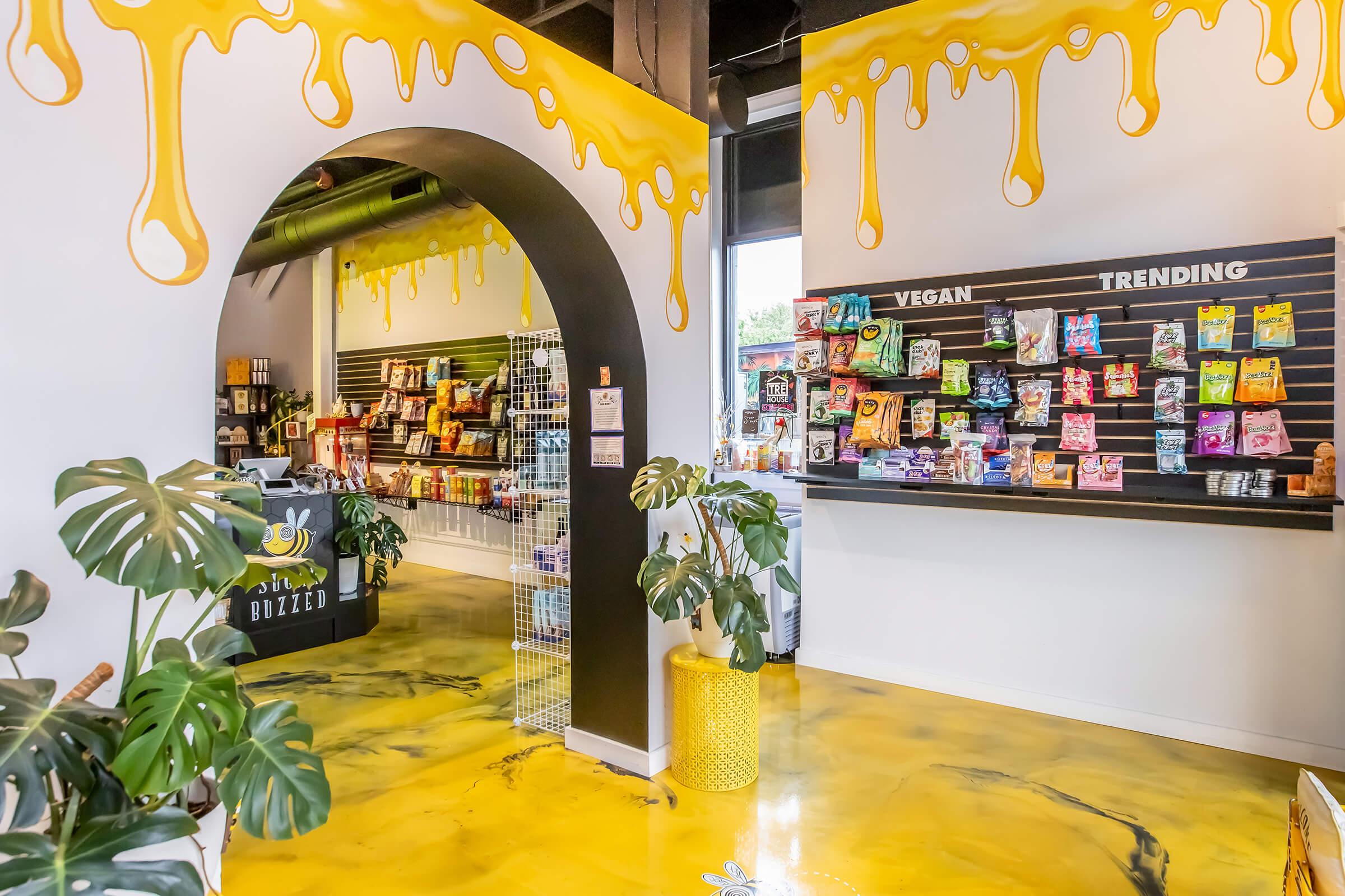Interior of a colorful shop featuring vibrant yellow dripping paint on the walls. An archway leads to a merchandise area, displaying various snacks and vegan products on shelves. Decorative indoor plants add a fresh touch to the modern space, designed with a bright, inviting atmosphere.