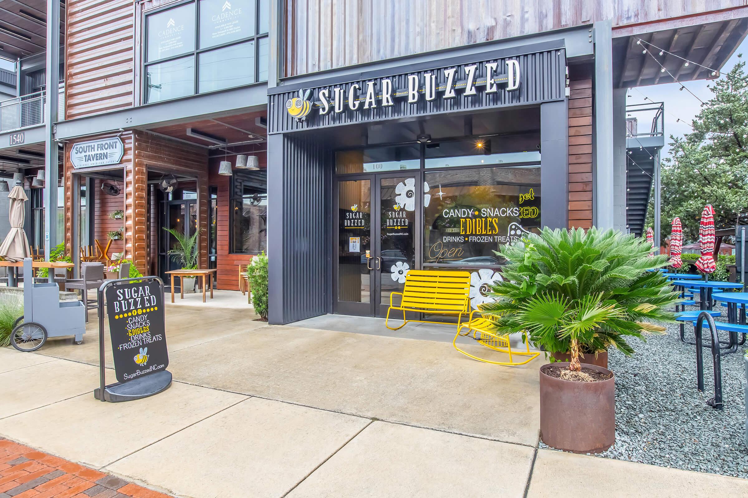 Exterior view of "Sugar Buzzed," a candy and snack shop with a bright sign. The storefront features large windows and a welcoming entrance, adorned with a yellow bench and tropical plants. The surrounding area includes tables and chairs, creating an inviting atmosphere for customers.