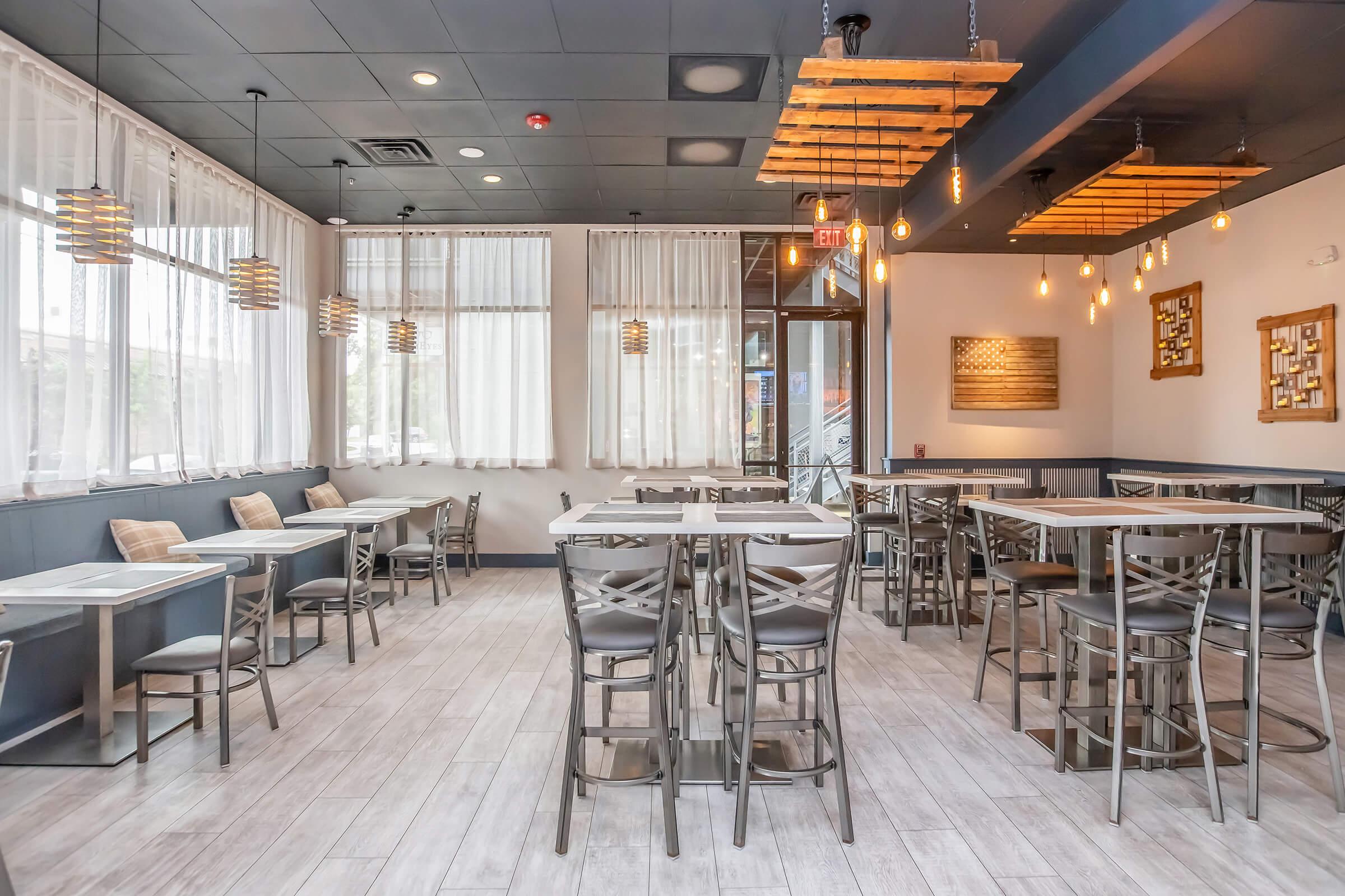 Interior of a contemporary restaurant featuring several dining tables and chairs, pendant lighting, and large windows with sheer curtains. The space is well-lit, creating a warm atmosphere, with wooden accents and a modern design aesthetic.