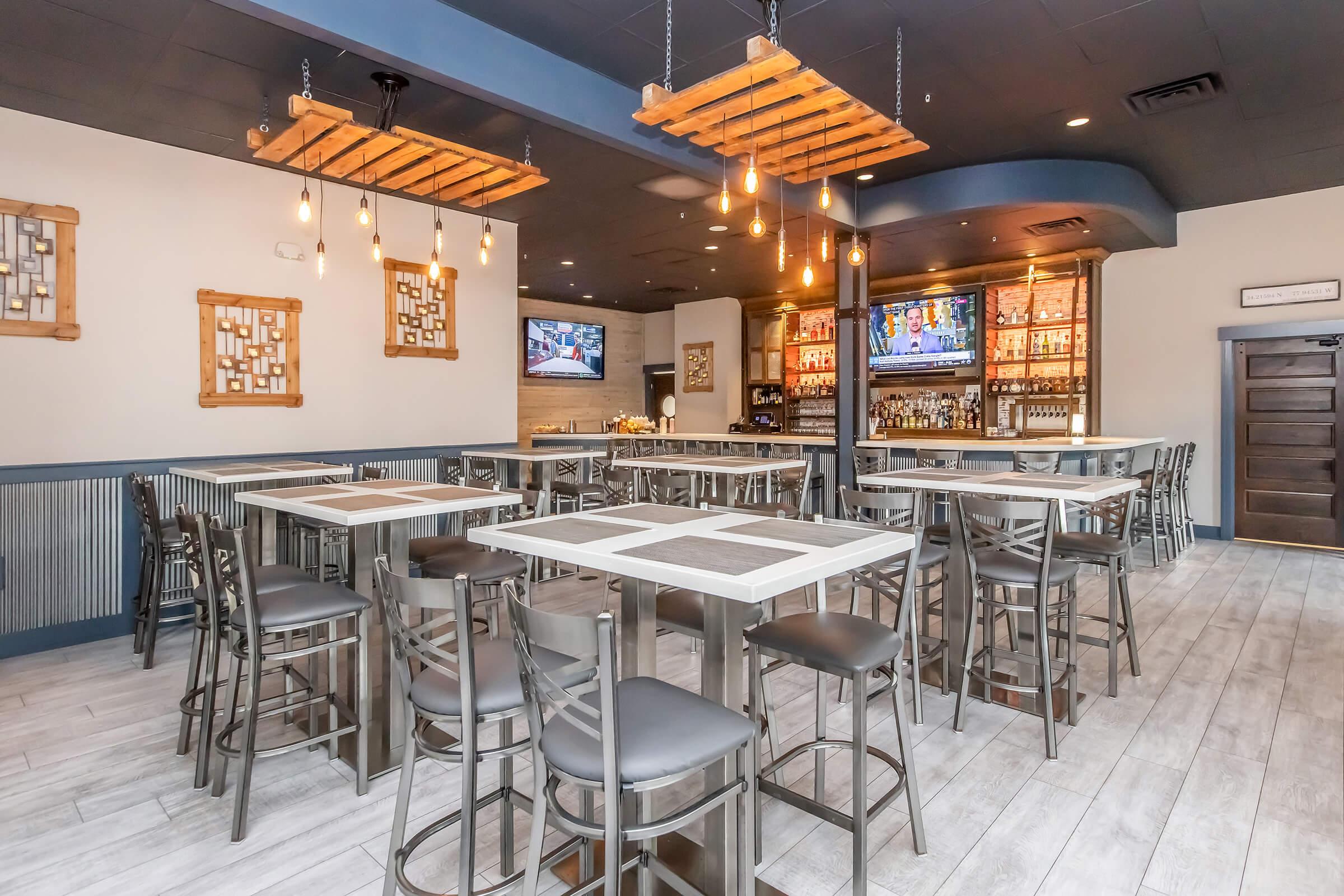 A modern dining area featuring wooden tables and high-backed chairs arranged neatly. The space is illuminated by pendant lights, with decorative wooden elements on the ceiling. A bar area with shelves displaying various beverages is visible in the background, along with two large televisions mounted on the walls.