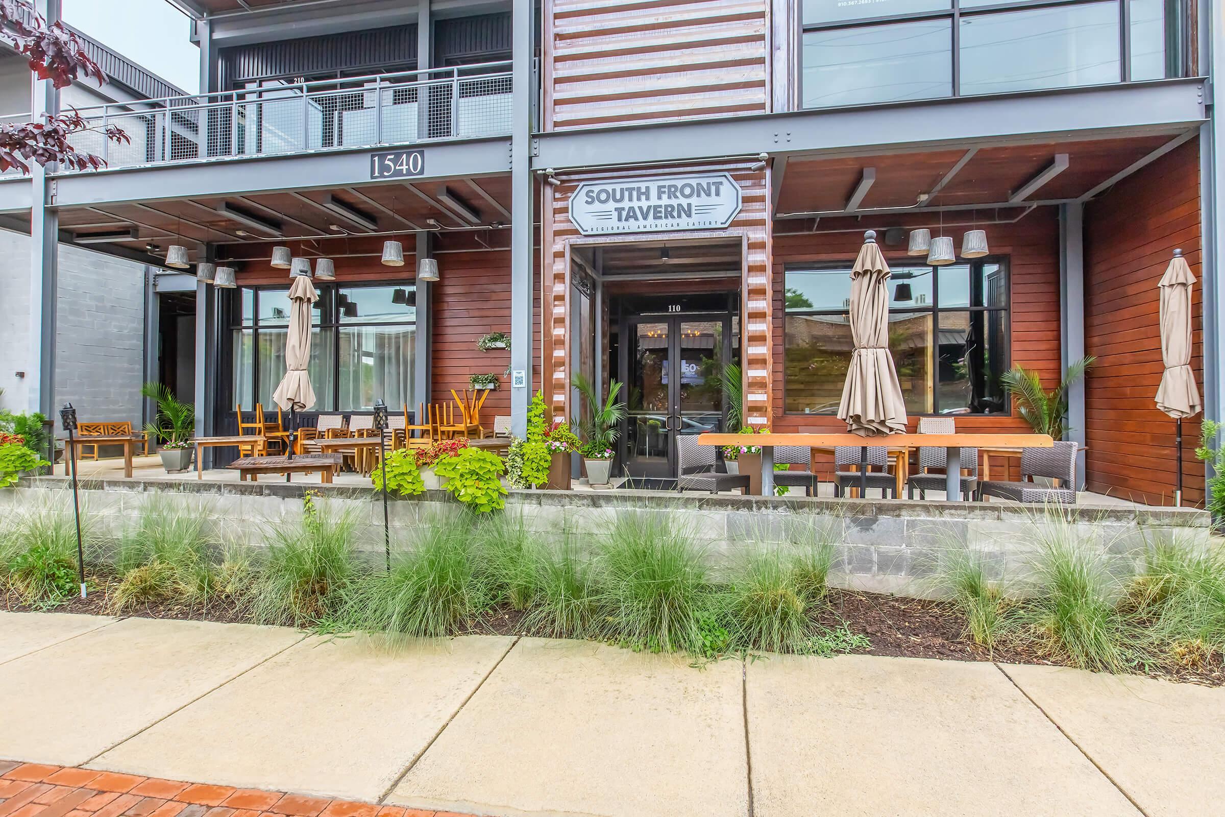 Exterior view of South Front Tavern, featuring a modern building with wooden accents, outdoor seating with tables and umbrellas, and landscaping with plants along the walkway. The entrance has glass doors and a sign displaying the tavern's name.