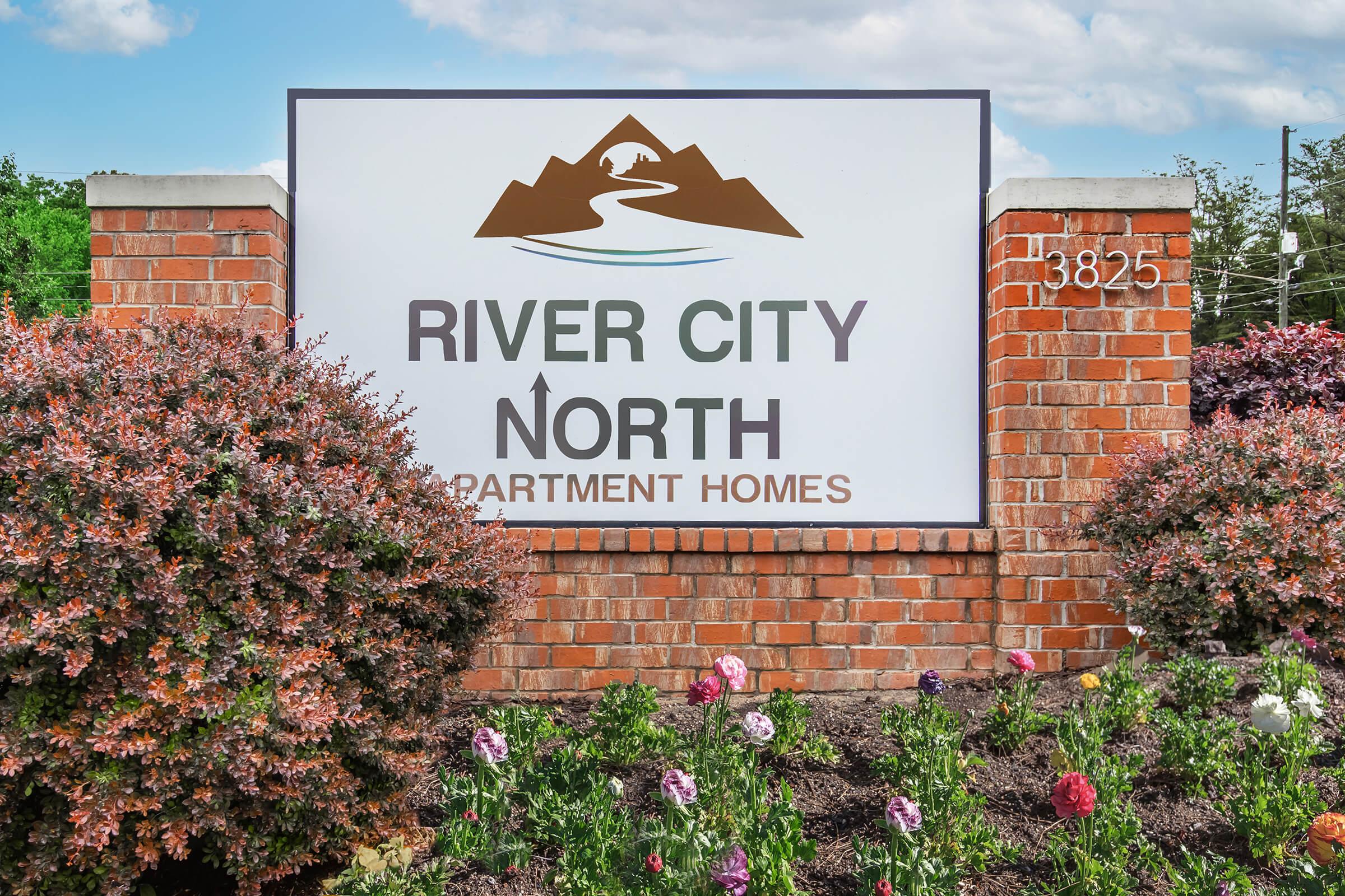 Sign for River City North Apartment Homes, featuring a logo with mountains and a river. The sign is set against a brick backdrop with colorful flower beds in the foreground. The address "3825" is displayed on the side. The sky is partly cloudy.